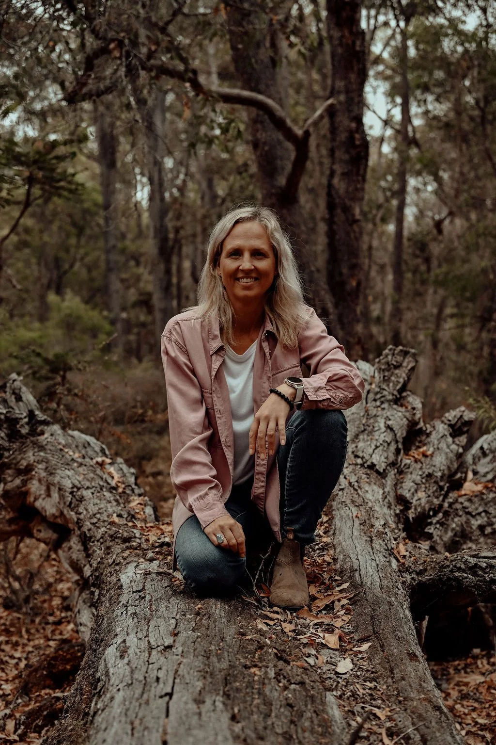 A woman with blonde hair smiling at the camera, kneeling on a fallen tree in a forest.