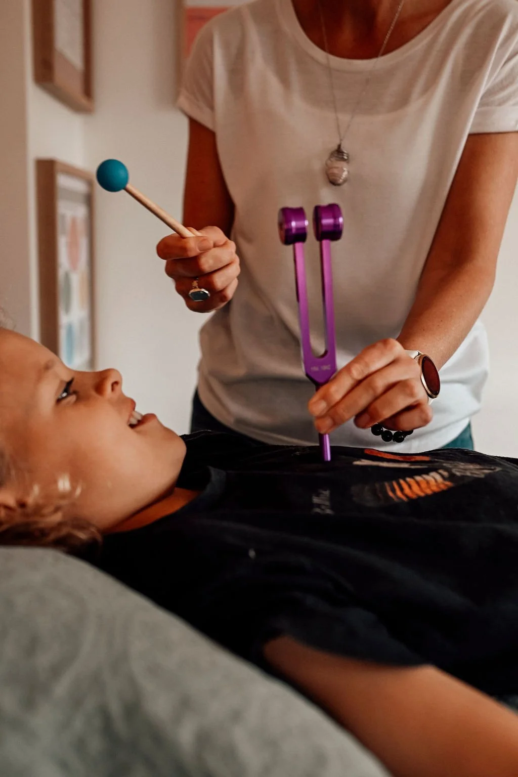 A young boy lying on a treatment table, having a kinesiology balance with a woman practitioner. They are using tuning forks to balance, which is held by the woman. The woman appears to be clearing the child with the tuning forks