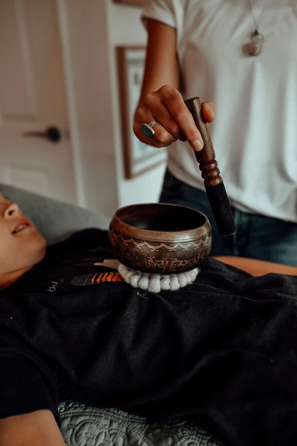 Person holding a wooden mallet over a singing bowl placed on a white padded cushion, with a child lying nearby in a relaxed setting.