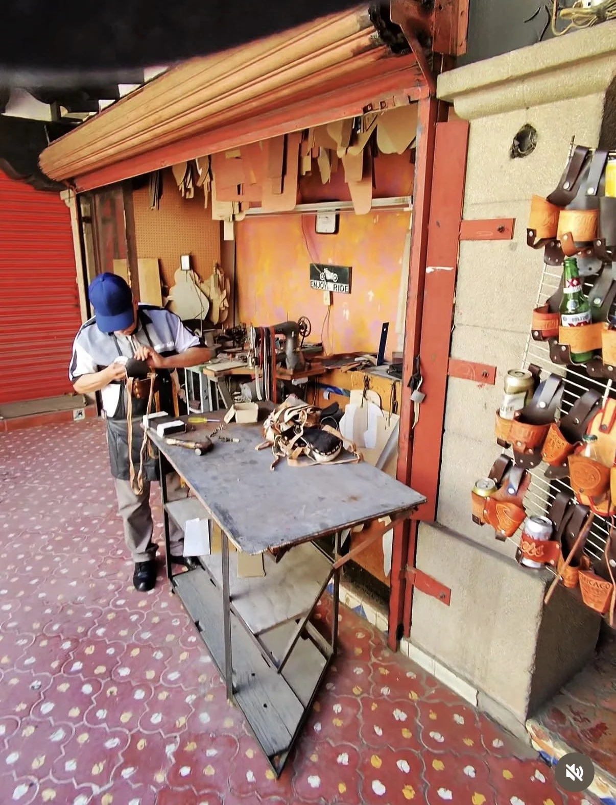 A man working in a small workshop with tools, on a workbench with leather straps, and a display with small orange containers holding tools and supplies. The workshop has a colorful, floral-patterned floor and a red wall with hooks and shelves.