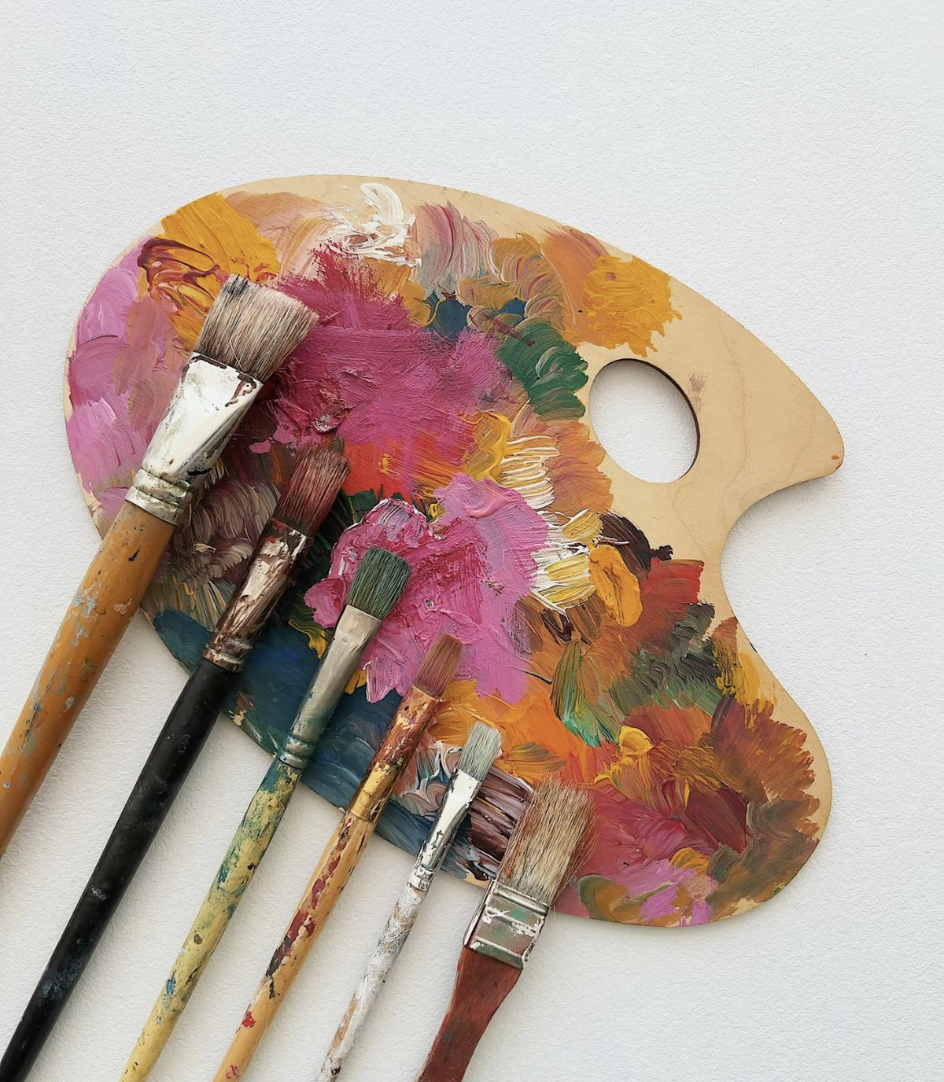 A wooden artist's palette with colorful paint blobs and six paintbrushes resting on it, against a plain white background.