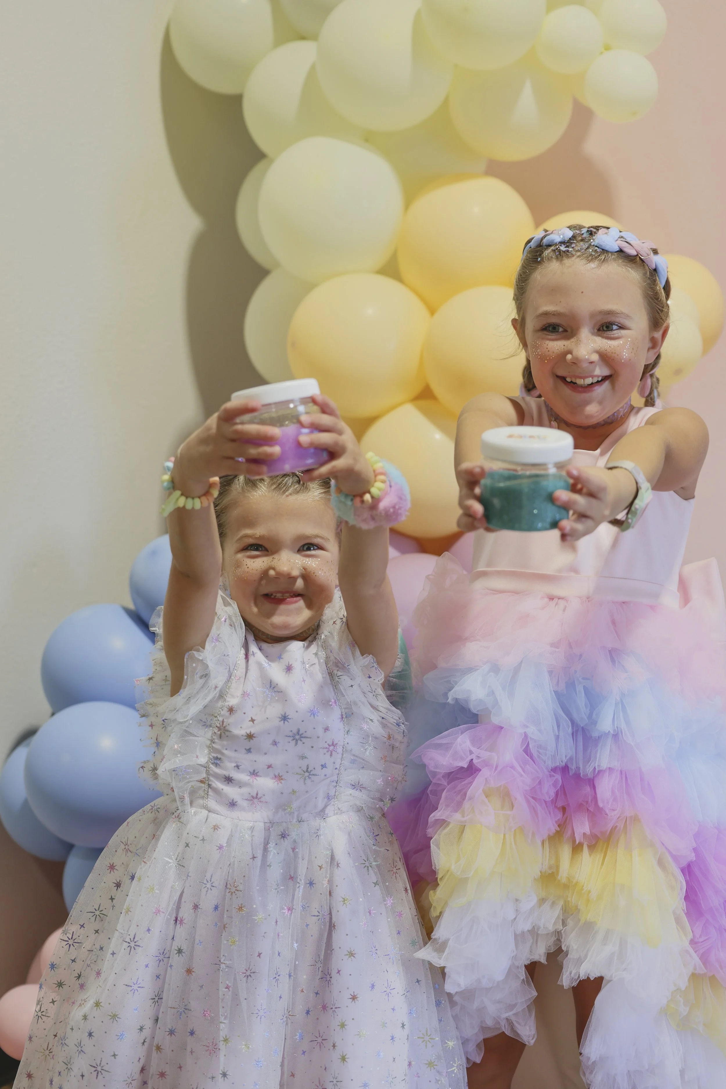 Two young girls in fairy-tale costumes smiling and holding jars of glitter, standing in front of a backdrop of pastel-colored balloons, with a girl dressed as a fairy in a multicolored tutu.