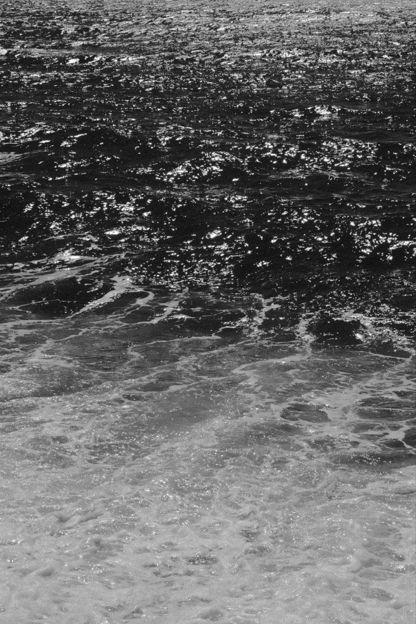 Close-up of ocean waves reflecting sunlight, with the water appearing dark in the background and foam at the shore in the foreground.