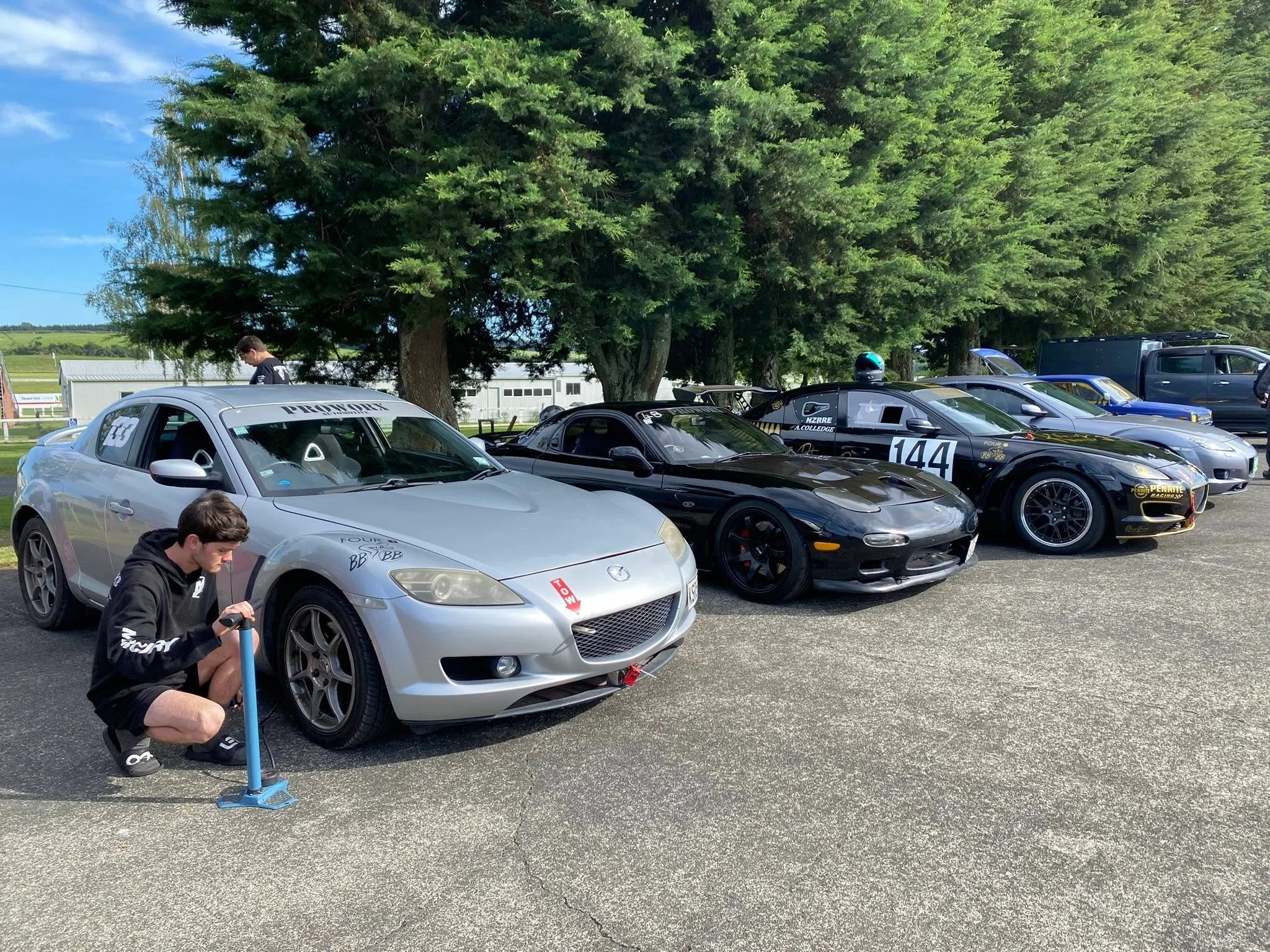 A parking lot with several race cars and a van parked under large green trees, with people working on the cars and preparing for a race.