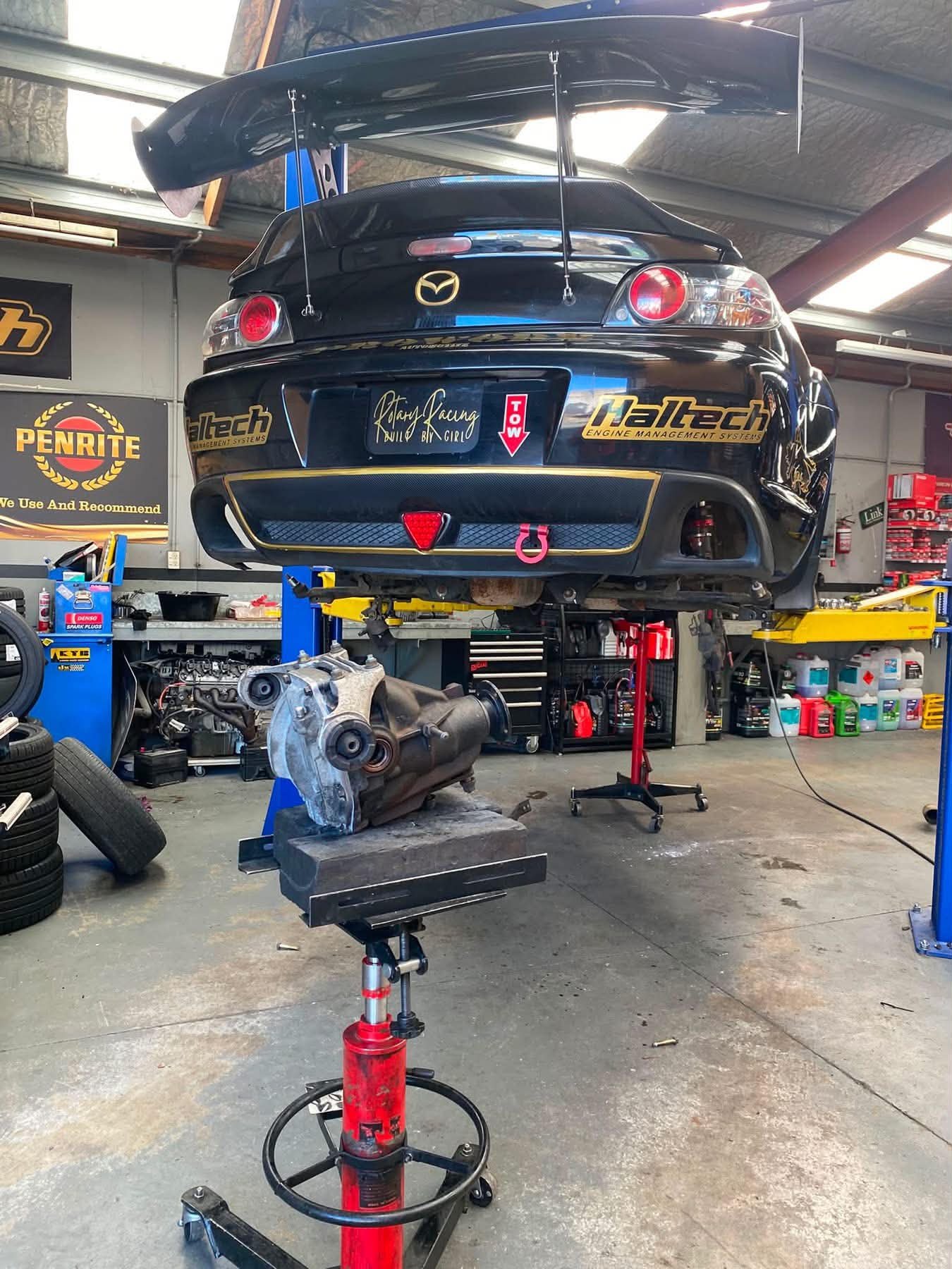 A black Mazda sports car with a large rear wing is elevated on a lift in an automotive workshop. The workshop has various car parts, tools, and containers in the background, along with a sign that says 'Penrite'. An engine block is placed on a stand below the car, surrounded by tires, and the garage has a concrete floor and metal ceiling.