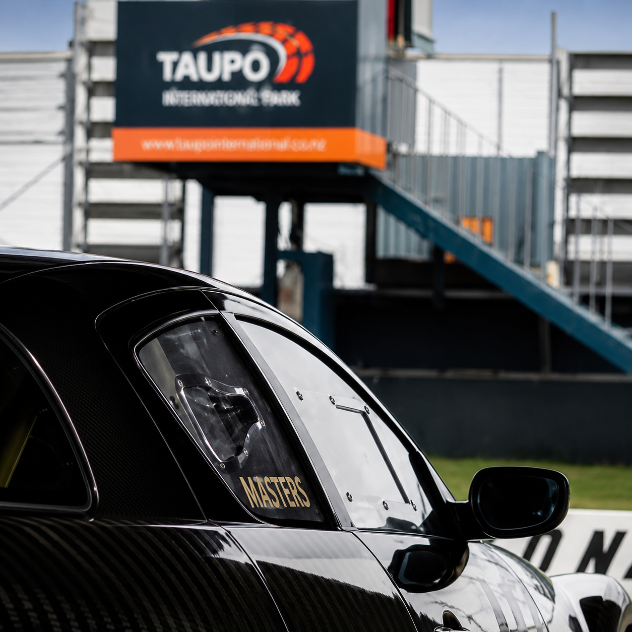 Close-up of a black race car with the word "Masters" on the window, at Taupo International Park with a grandstand and signage in the background.