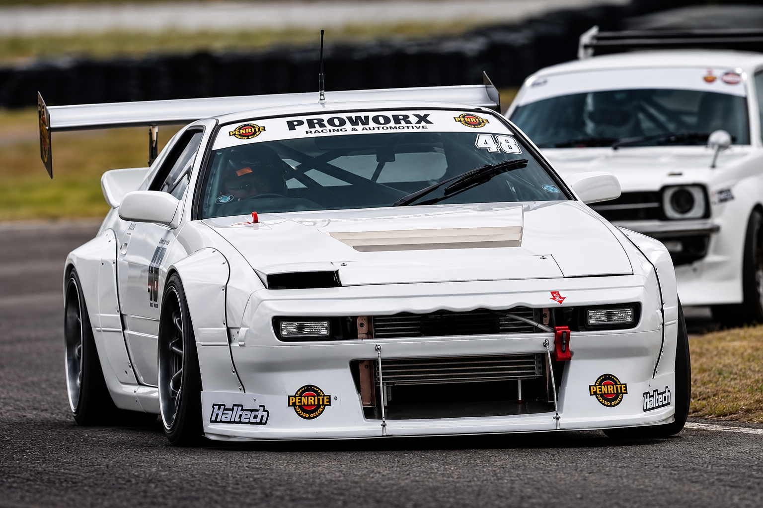 A white race car with the number 48 on the windshield, parked on a race track, with another white race car in the background.