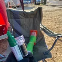A camping chair with a black backrest and armrests, with various camping supplies including bottles and containers resting on it, outdoors on a dry grassy area.