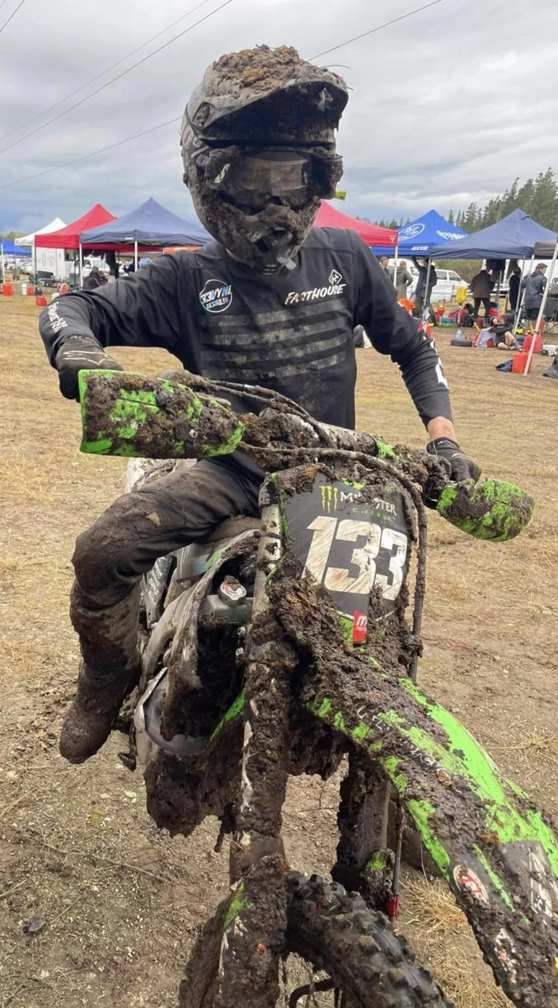 A dirt-covered motocross rider on a green motorcycle at an outdoor race track with tents in the background.