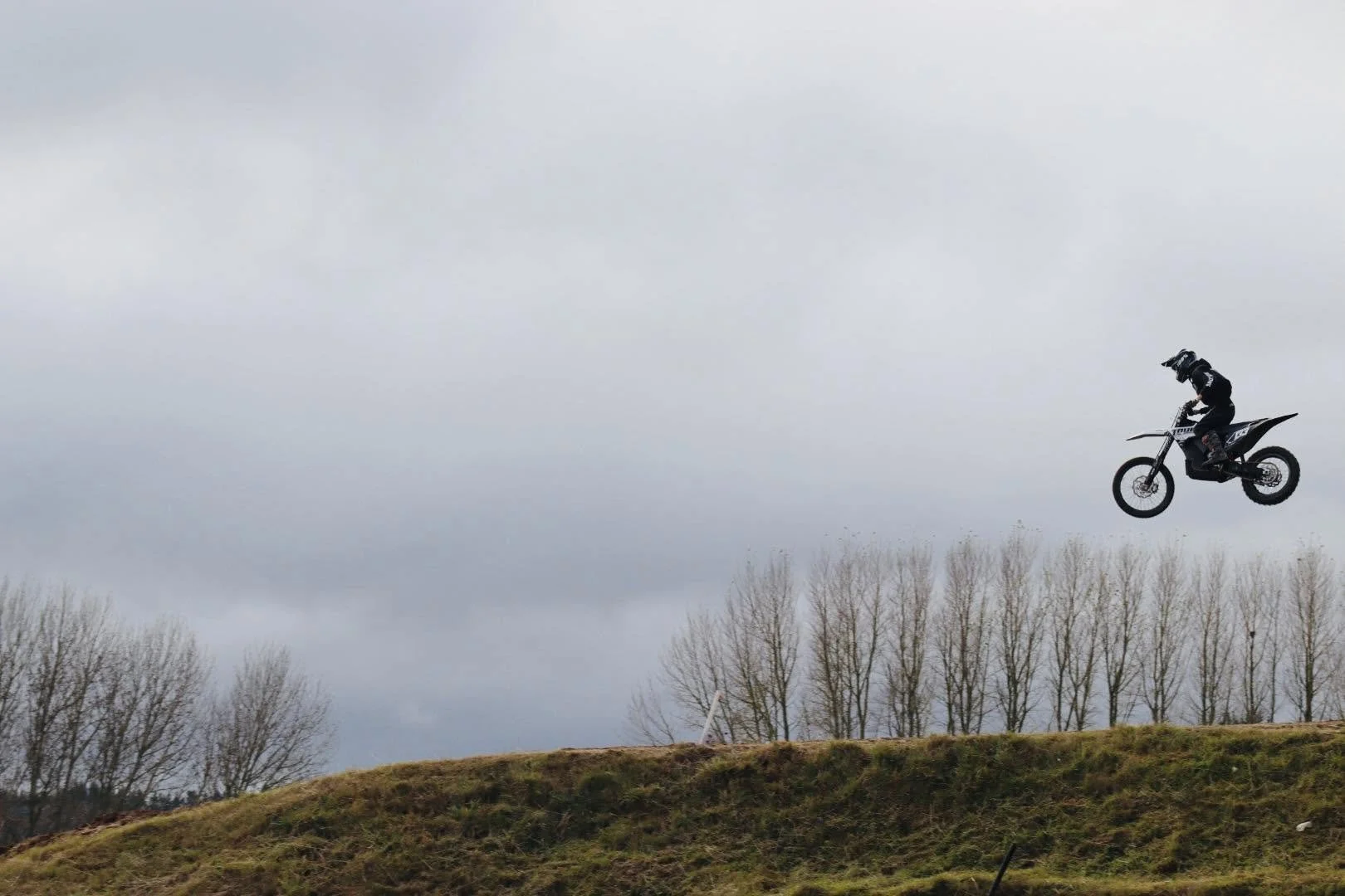 A person riding a motocross dirt bike in mid-air against a cloudy sky background, with a line of bare trees on the horizon.