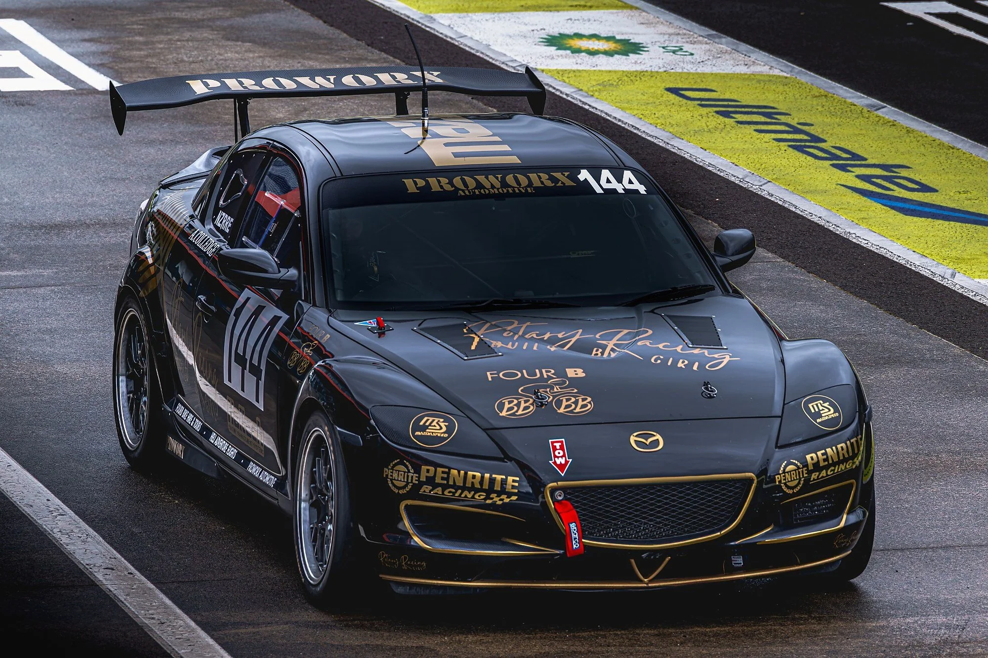 A black race car with racing logos and decals, including the number 144, parked in a pit lane with a yellow and blue caution sign on the ground.