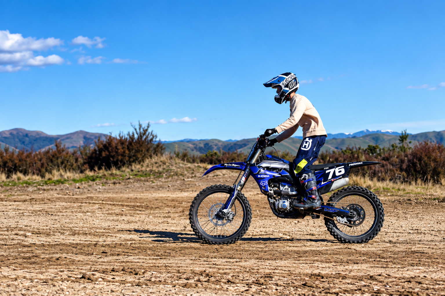 A person riding a blue dirt bike on a dirt trail with mountains and a clear blue sky in the background.