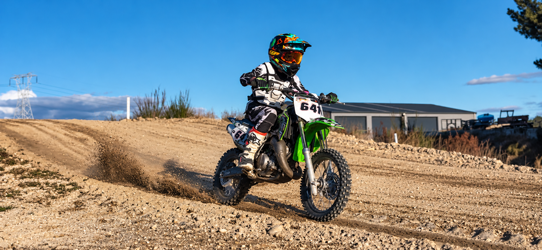 A child riding a green dirt bike on a dirt track, wearing full motocross gear and helmet, with a clear blue sky and a building in the background.