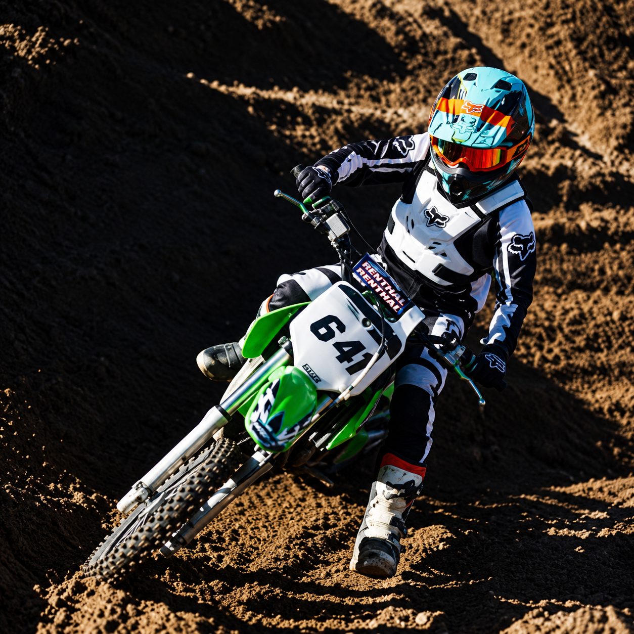 Motocross rider wearing a white and black suit and a colorful helmet, riding a green dirt bike on a dirt track.