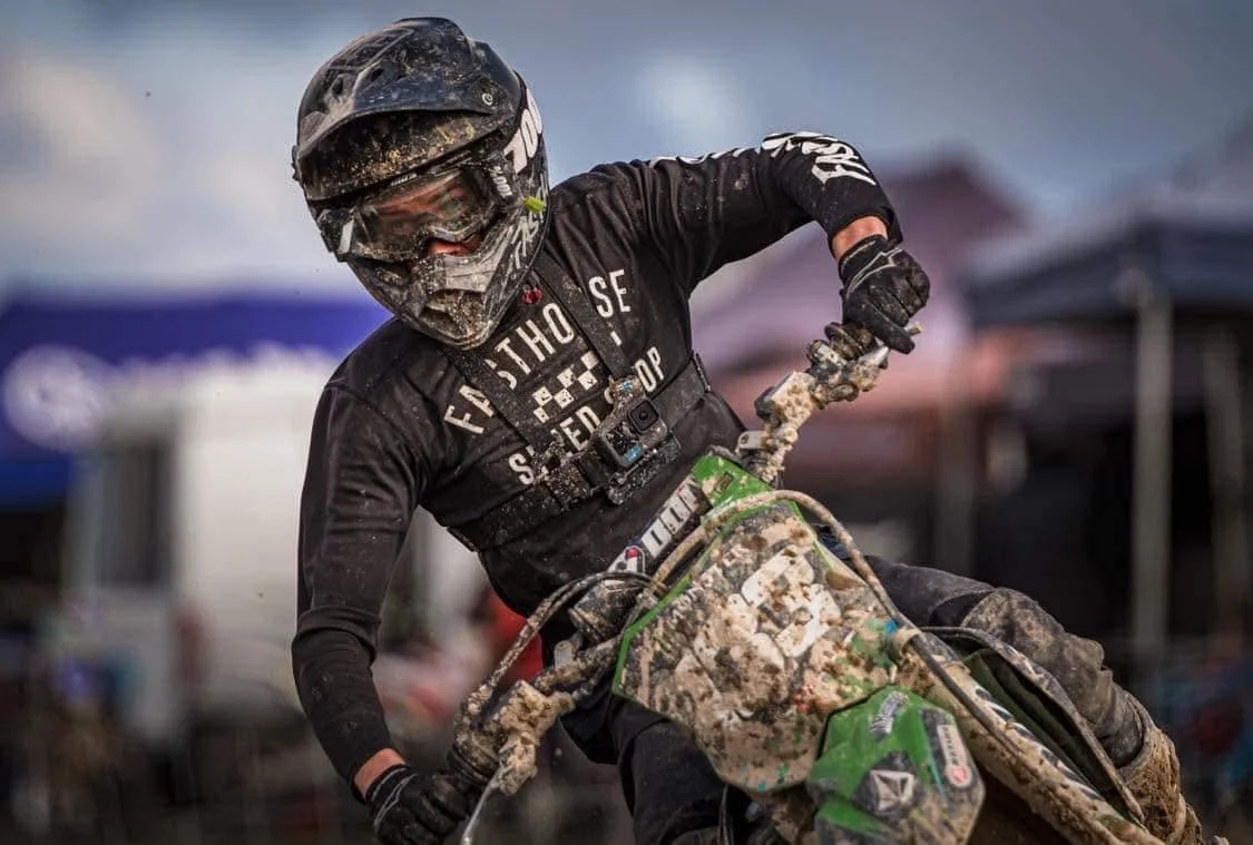 A motocross rider wearing a black helmet and gear, riding a muddy green dirt bike.