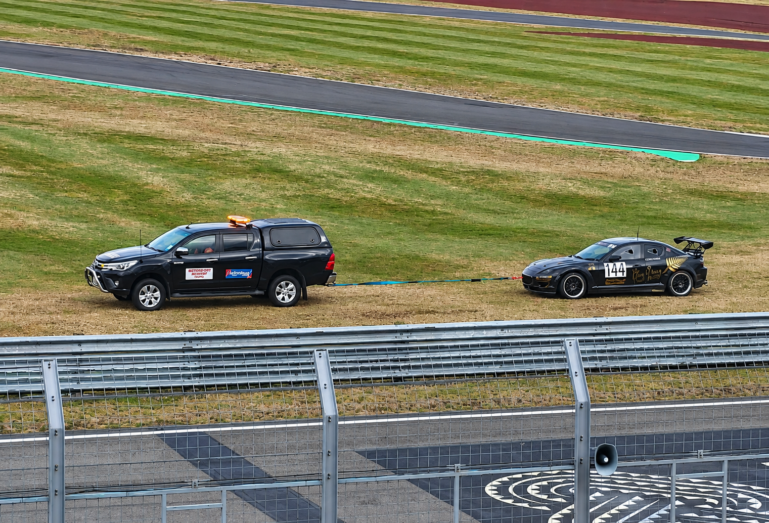 A black sports car with racing decals and the number 144 on the side is being towed by a black pickup truck with emergency vehicle markings on a grassy area near a race track.