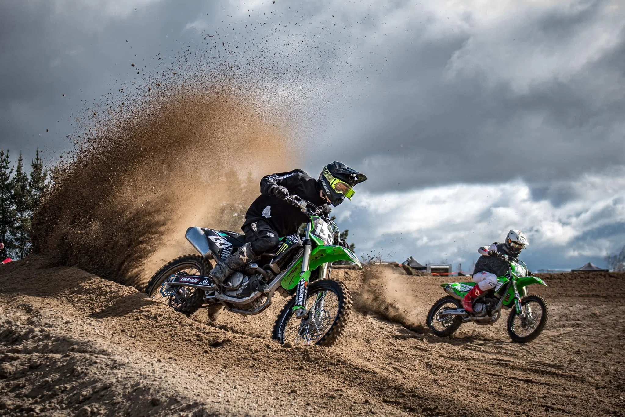 Motocross racers navigating a dirt track with one rider kicking up dust and gravel under a cloudy sky.