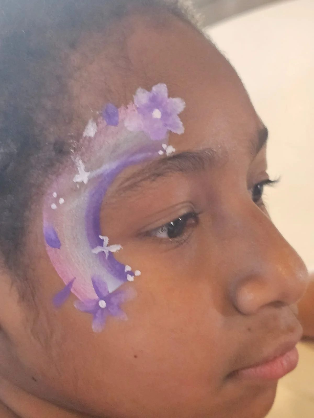 A close-up of a young girl's face with face paint featuring purple and pink flowers and white accents on her forehead and near her eye.