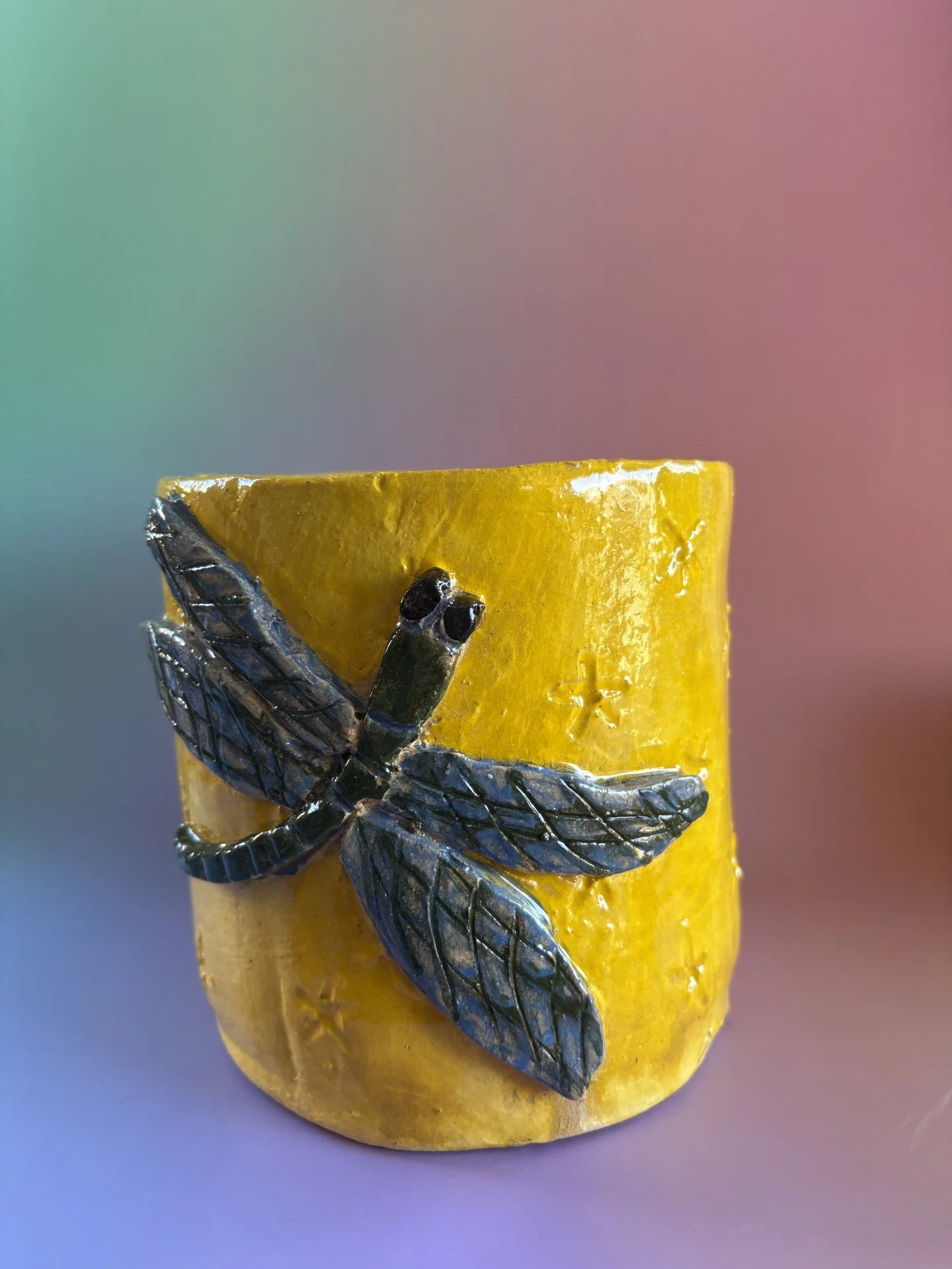 Yellow pot with a blue and green dragonfly decoration on the side, against a blurred pastel-colored background.