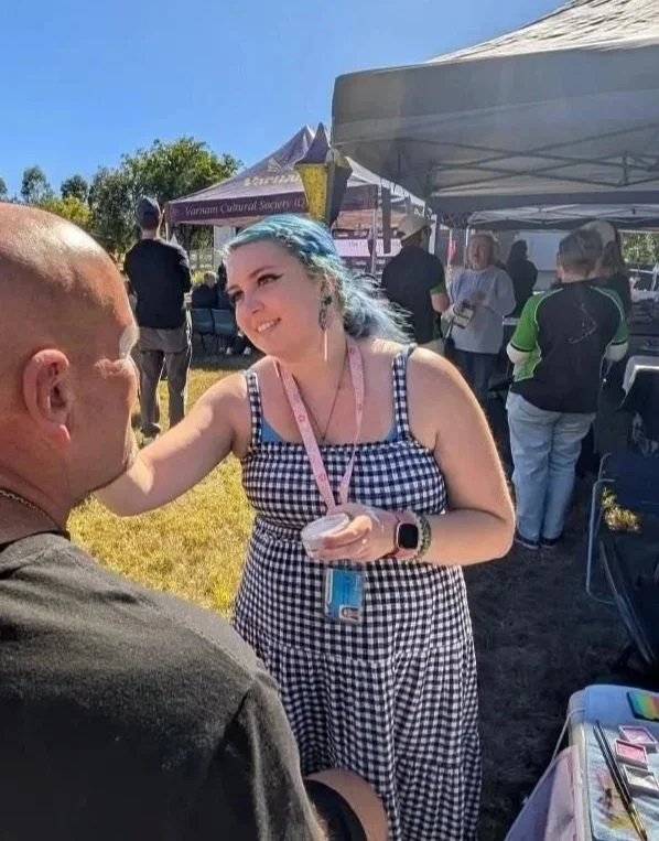 A young woman with blue hair, wearing a black-and-white checkered dress, smiling as she paints a man's face at an outdoor event with tents and people in the background.