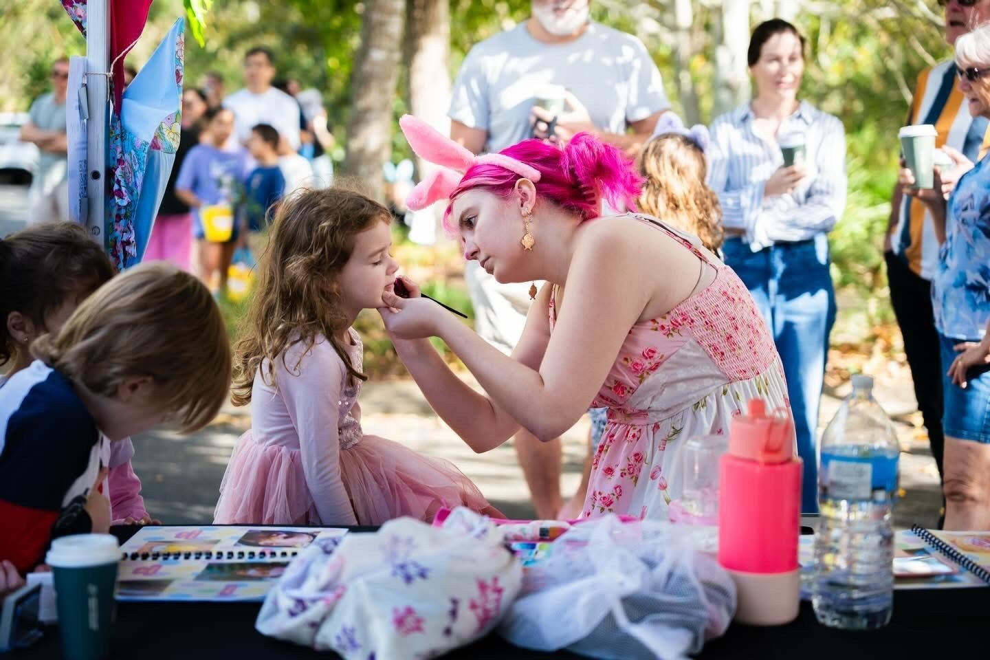 A young woman with bright pink hair, bunny ears, and gold earrings is painting a young girl’s face at an outdoor event during the day. The girl is wearing a pink tutu, and several people are watching in the background.