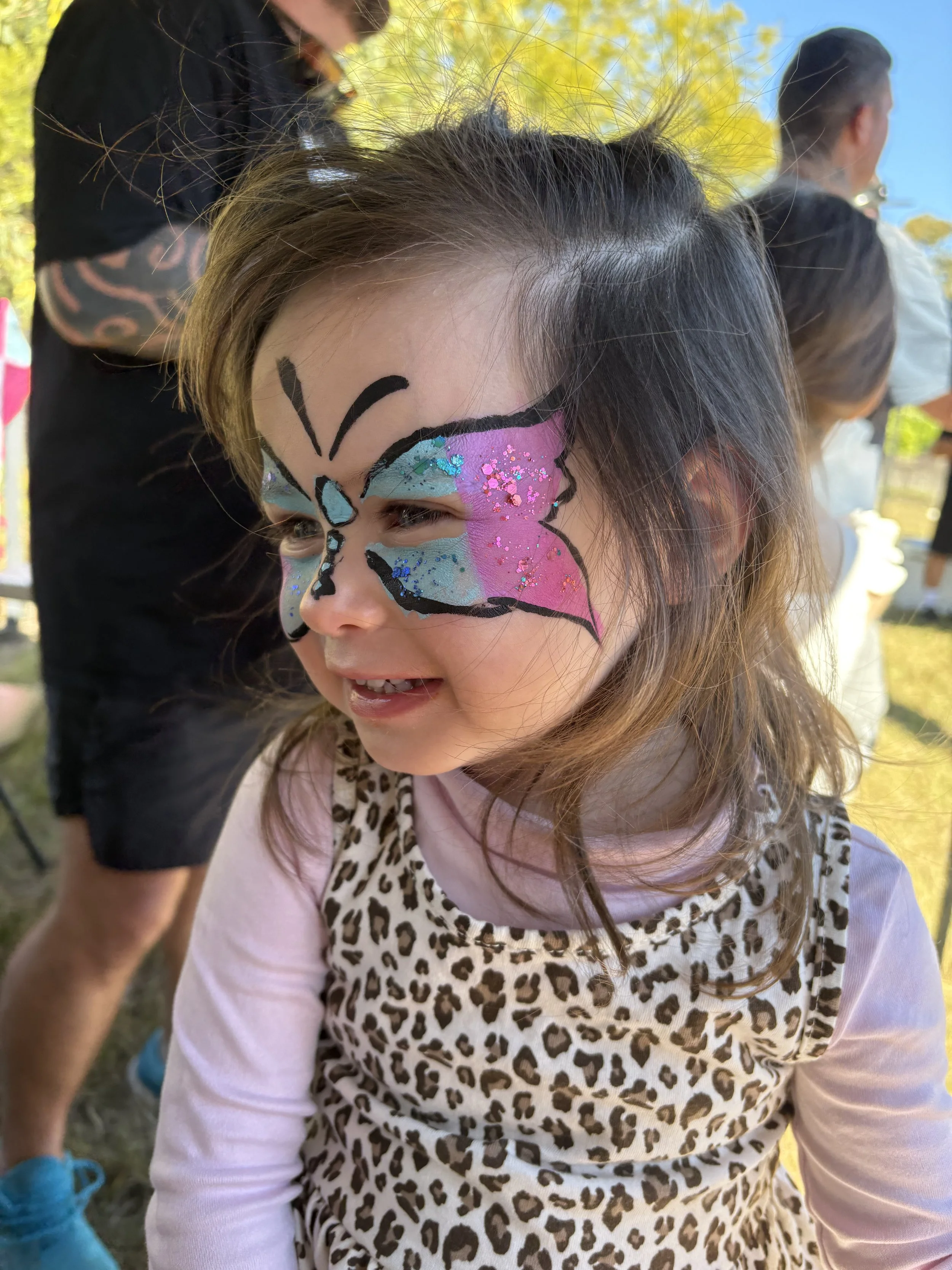 A young girl with face paint of a butterfly with pink, blue, and purple colours and glitter, wearing a leopard print dress, outdoors on a sunny day, surrounded by other children and adults.
