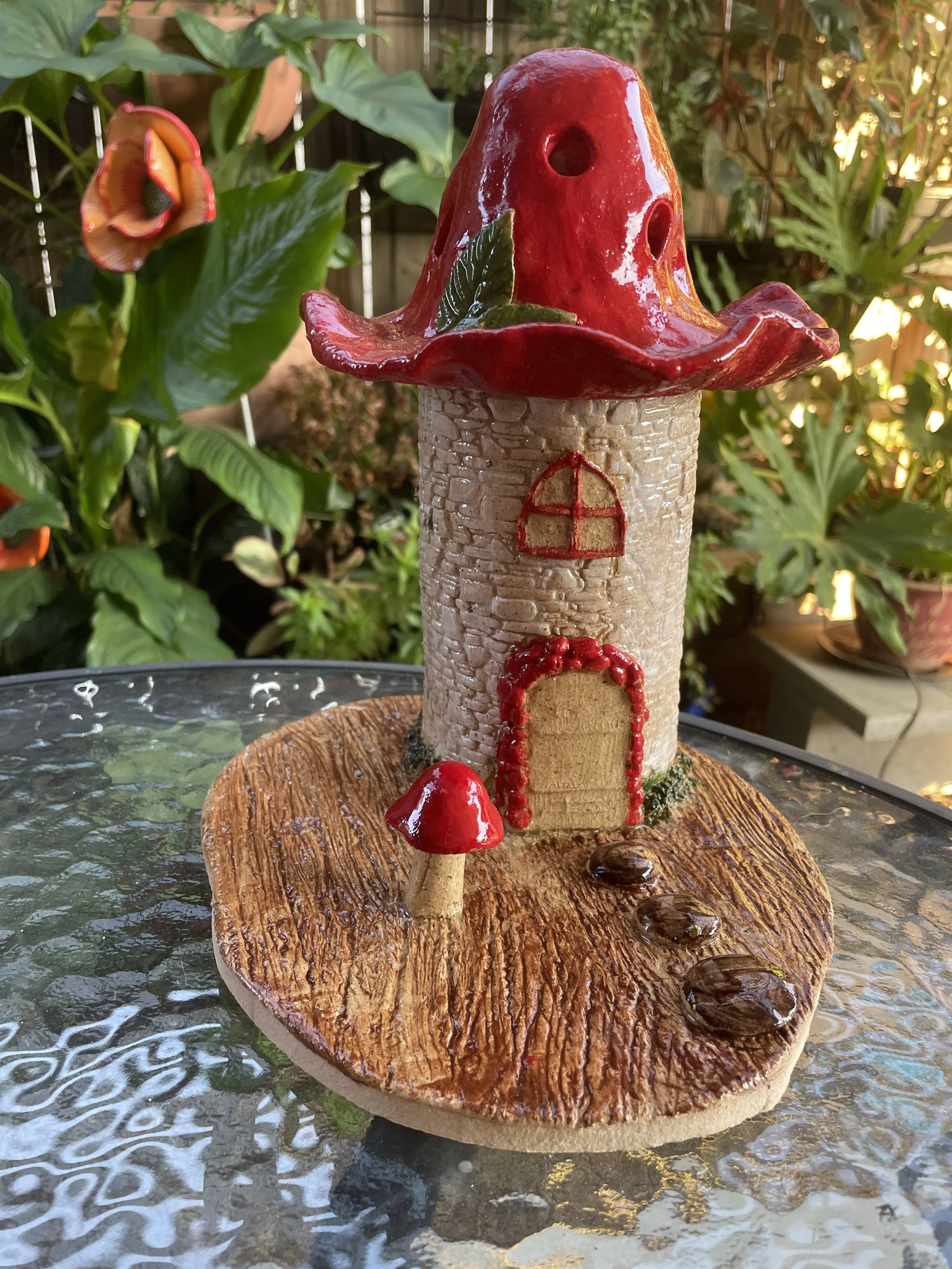A decorative small fairy tale house fountain with a red mushroom cap roof, a beige brick-like tower, red window and door accents, and red mushrooms at the base, set on a wooden platform in a garden surrounded by greenery.