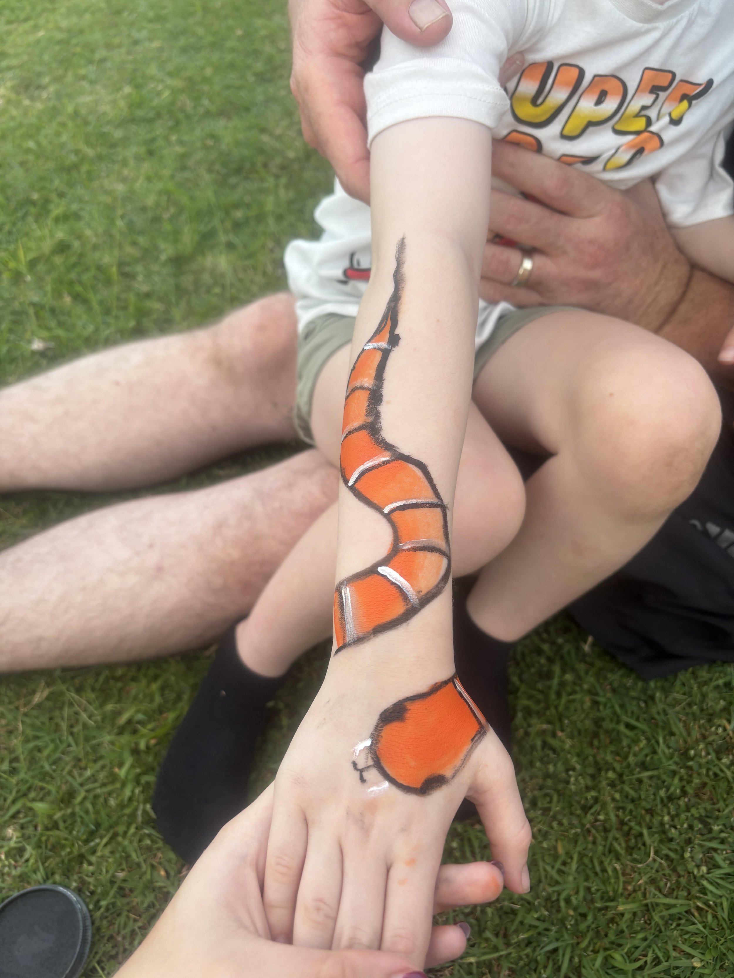 Child with a painted orange and black snake on their arm, sitting on grass.