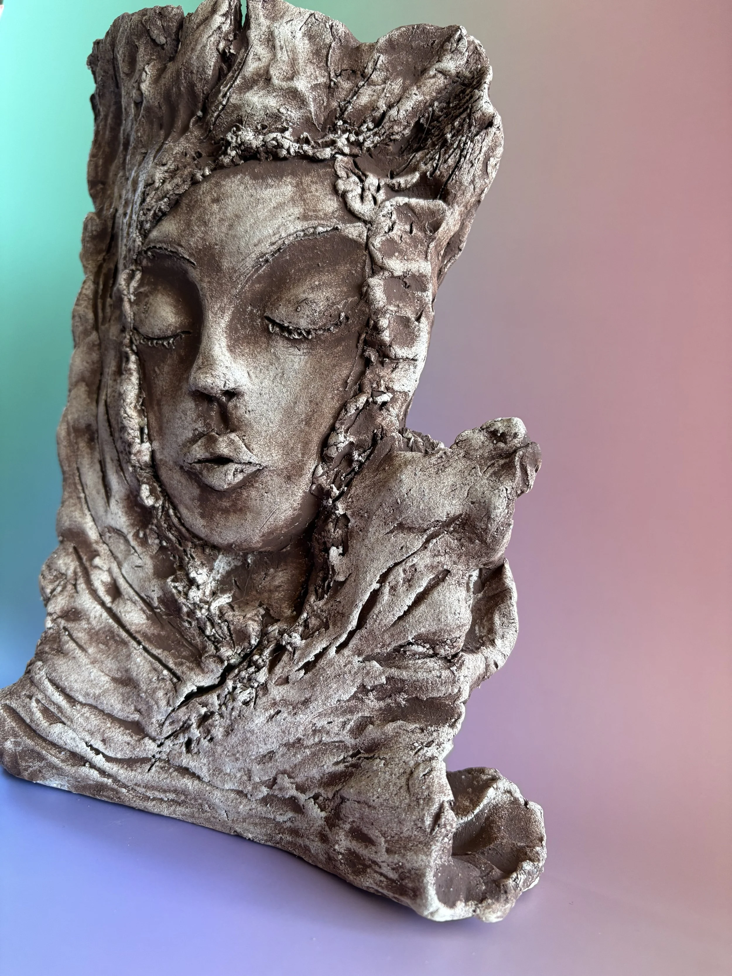 Close-up of a textured sculpture of a serene woman's face with closed eyes, resting her chin on her hand, made from clay and oxide, against a gradient pastel background.