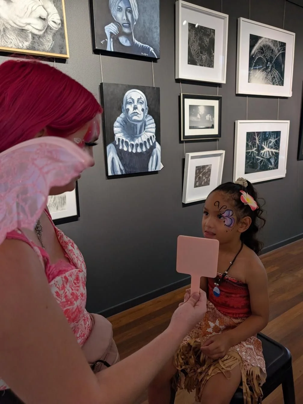 A girl with facepaint of a butterfly and flowers sits on a stool while a young woman with colourful hair paints her face at an art gallery. The gallery wall behind them displays framed artwork, including portraits, abstract images, and nature scenes.