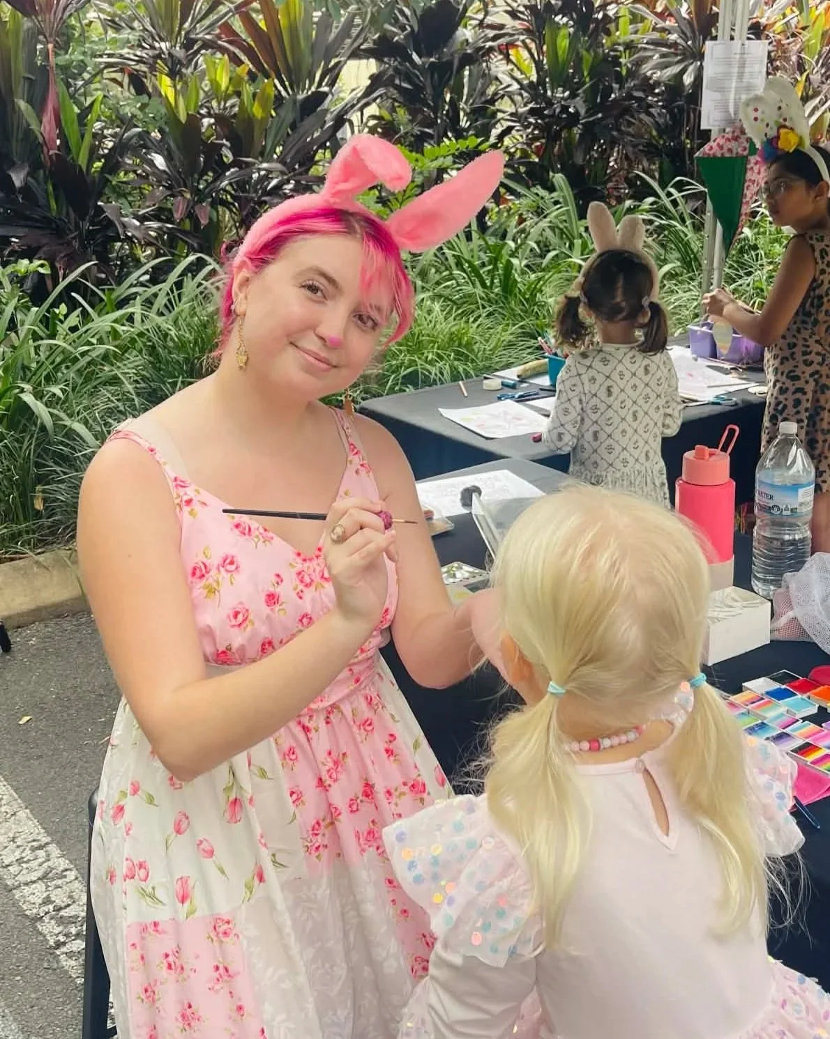 Young woman with pink hair and bunny ears, face painting a young girl with blonde hair, who is wearing a colorful beaded necklace and a pastel-colored dress, at an outdoor arts and crafts event.