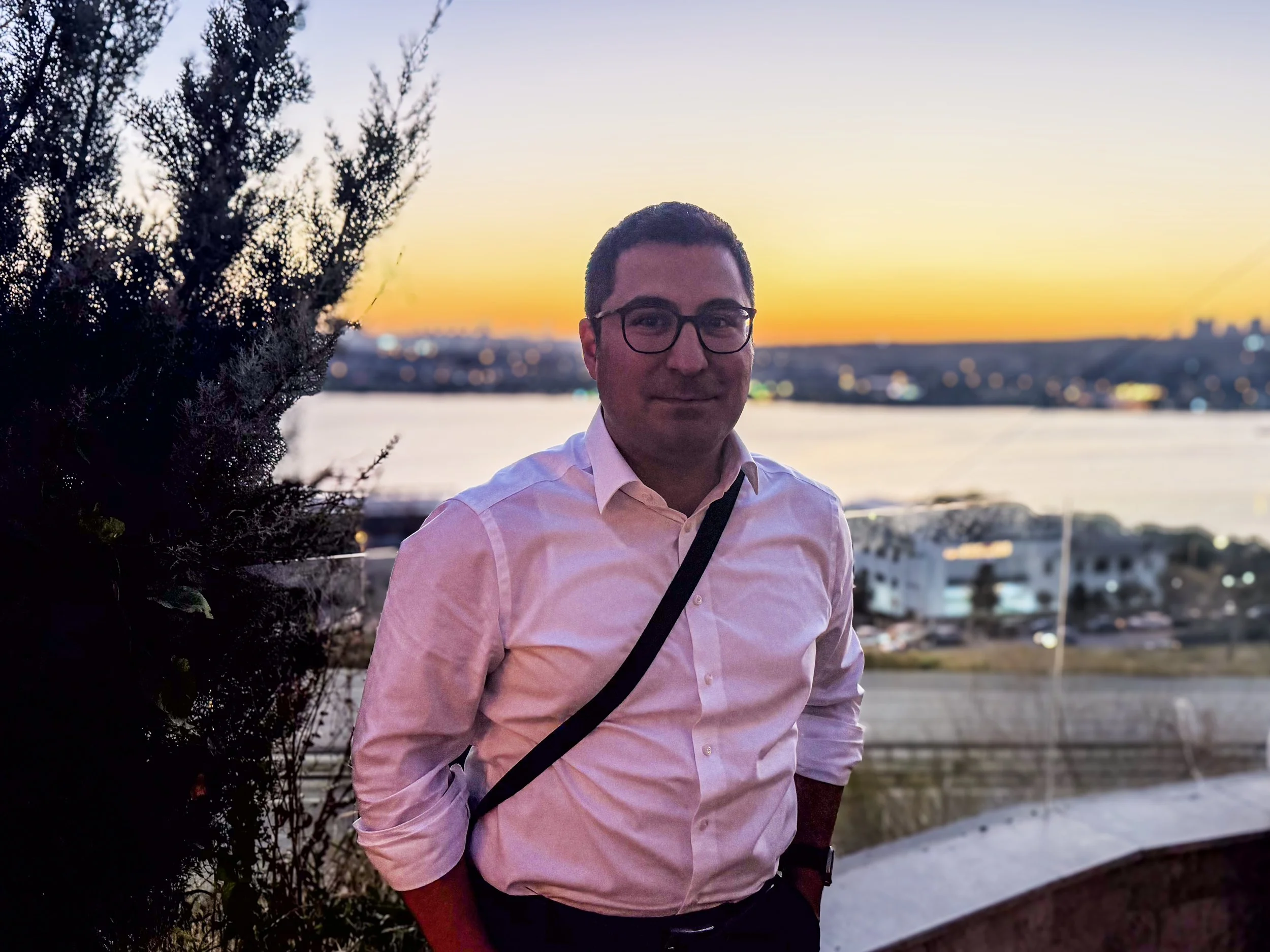 A man wearing glasses and a pink button-up shirt, standing outdoors during sunset, with a body of water and city lights in the background. He has dark hair and is smiling slightly.