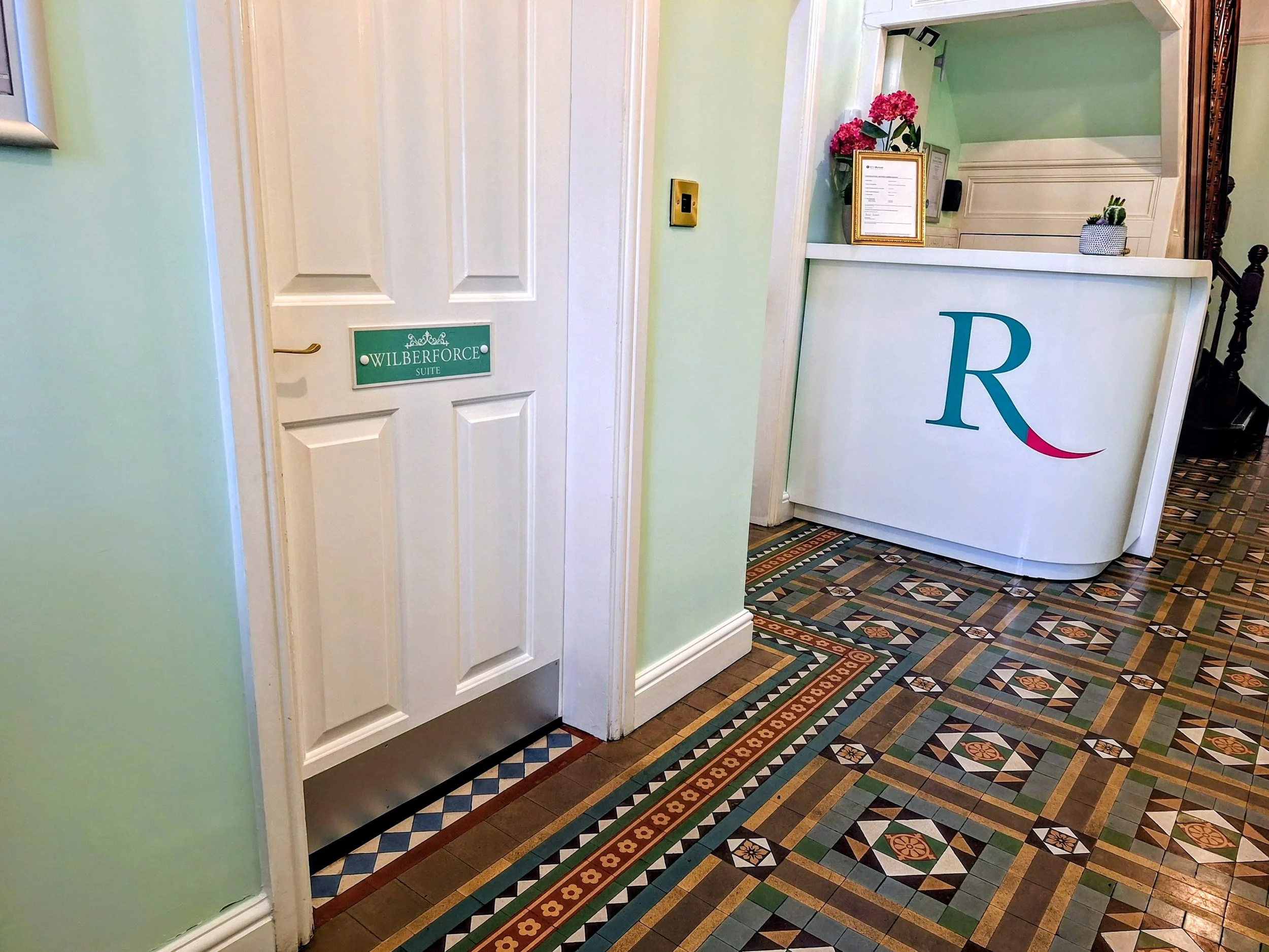 Interior of a building with patterned tile flooring, a white door labeled 'Wilberforce Suite,' and a reception desk with a large 'R' on the front, decorated with pink flowers and a small cactus.