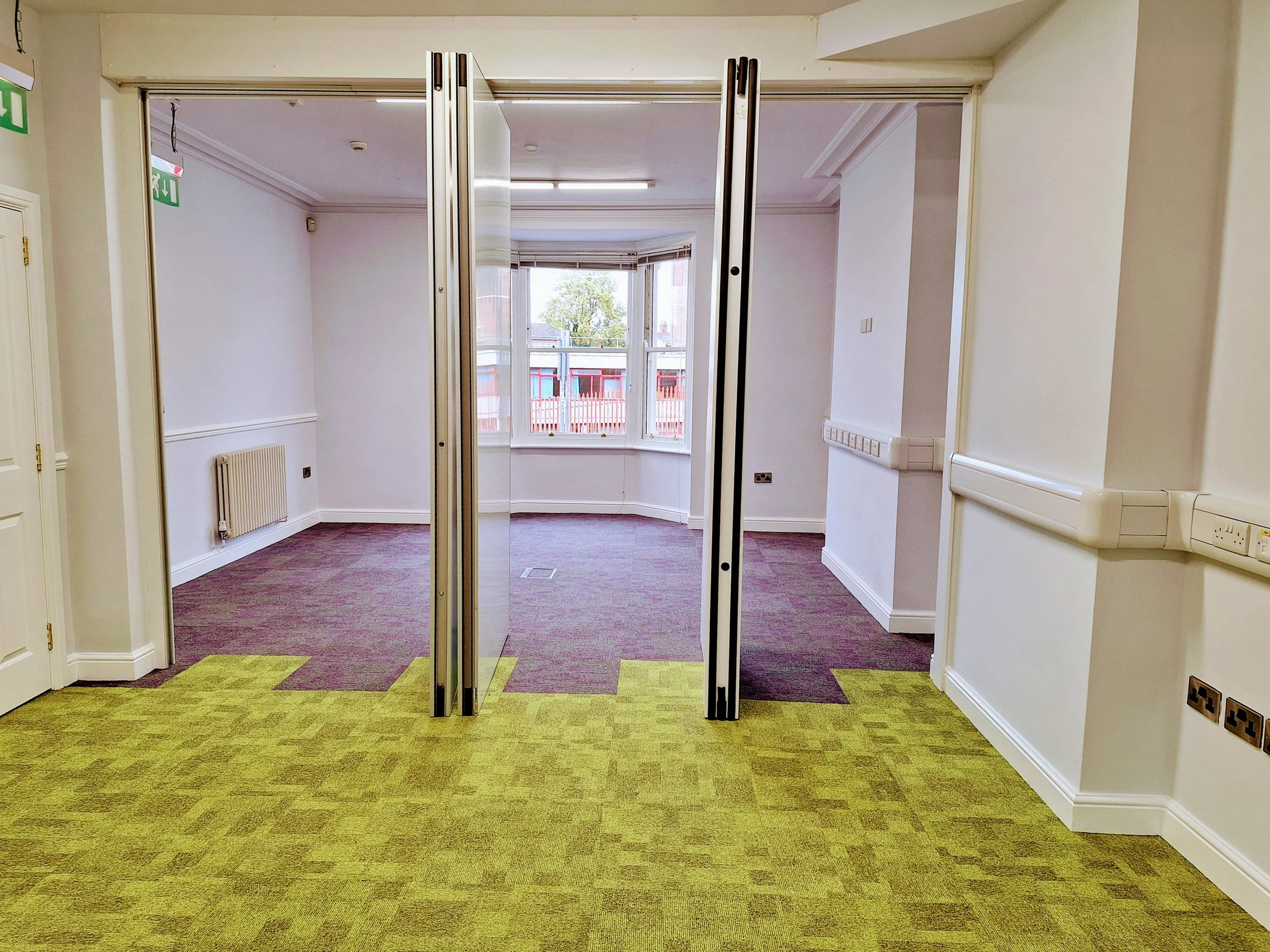 Empty carpeted office interior with folding glass doors, bay window, radiator, and wall-mounted electrical outlets