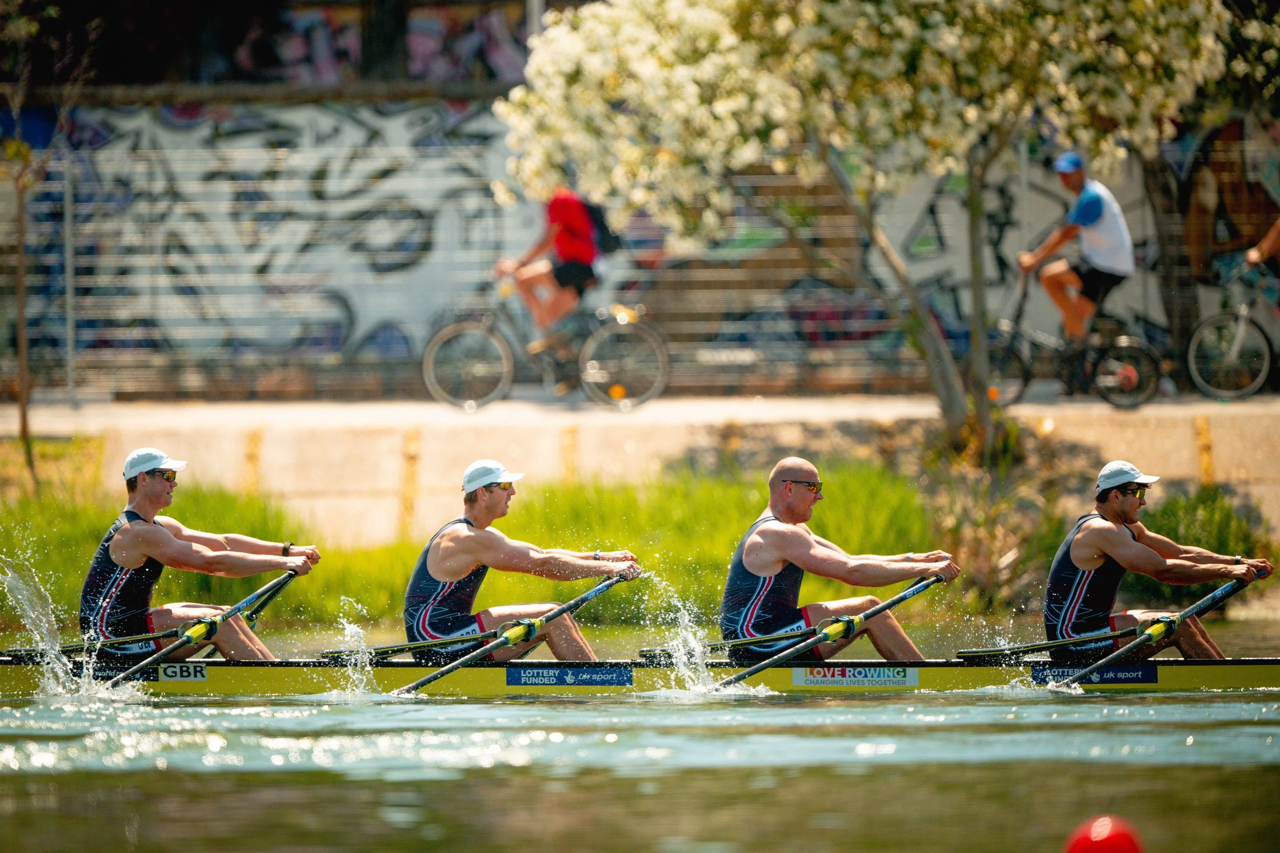 A group of male rowers racing in a boat on a river, pulling the oars in unison with water splashing around them, with spectators and a graffiti wall in the background.