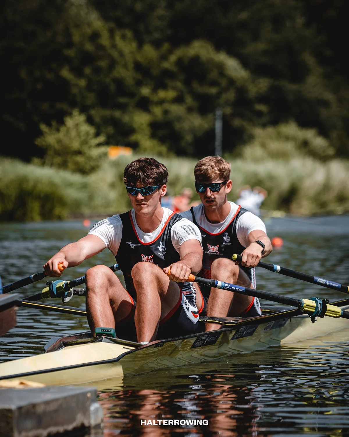 Two male rowers in a yellow boat competing in a rowing race on a river, with trees and spectators in the background.