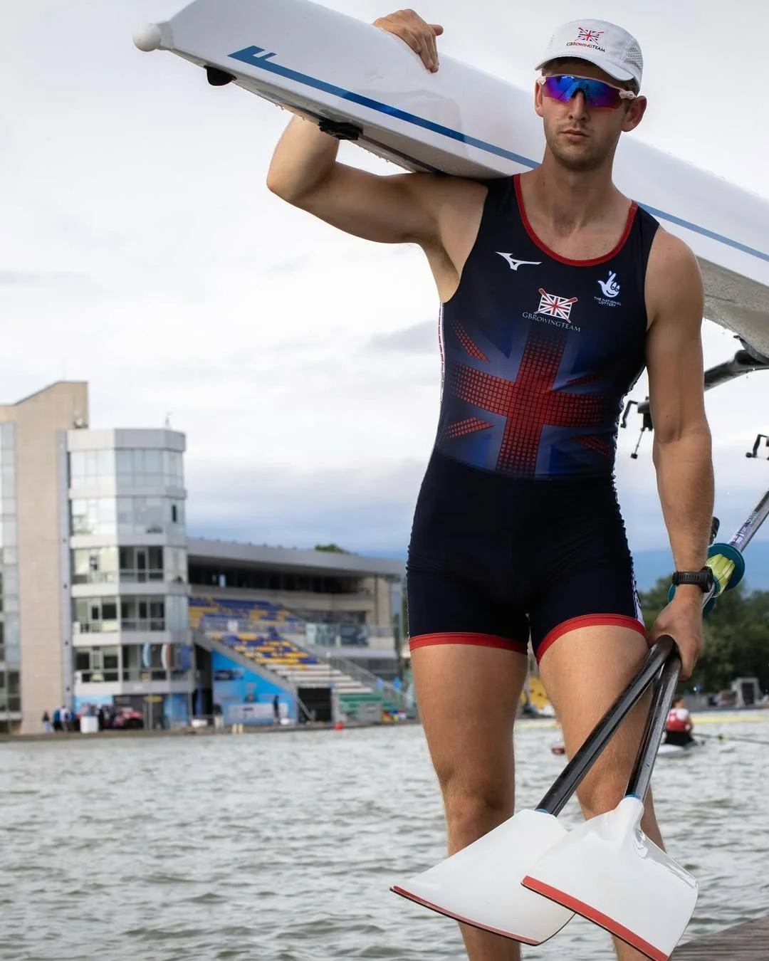 A male athlete in a triathlon uniform carrying a kayak paddle, standing by a river with a building and bleachers in the background.