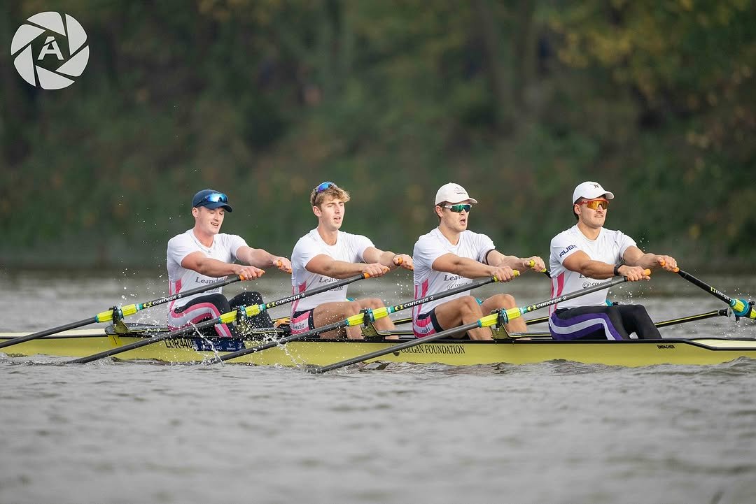 Four male rowers in a yellow boat on a lake, all wearing white shirts, racing caps, and sunglasses, with lush green trees in the background.