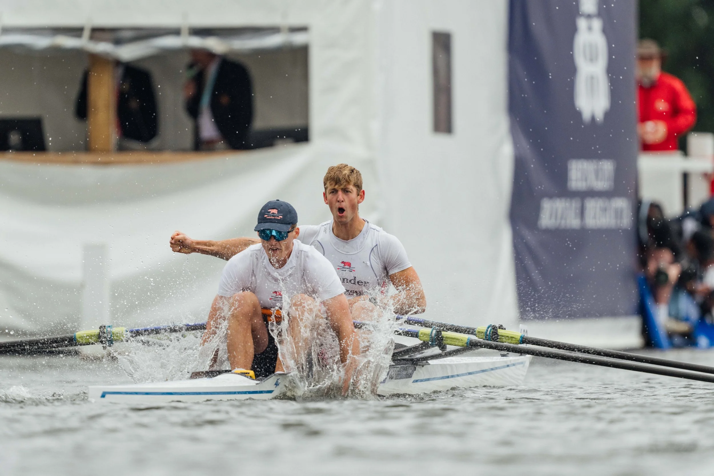 Two young male athletes paddling a dragon boat on a water body during a race, with spectators and banners in the background.
