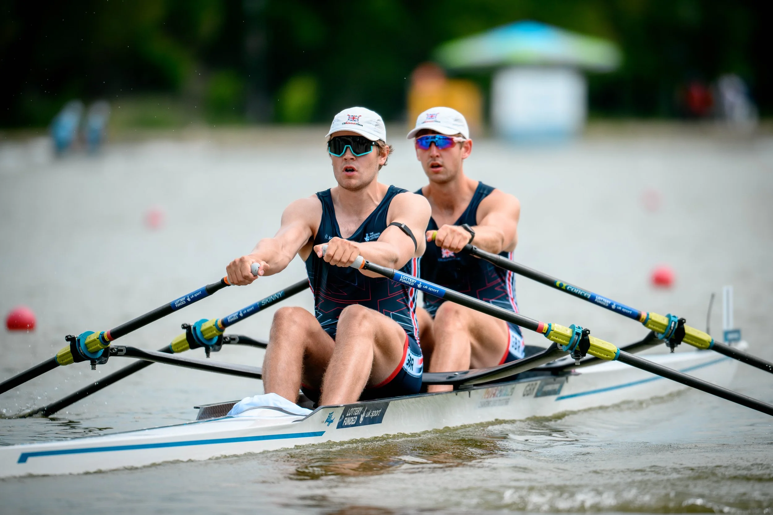 Two male athletes in a double sculls rowing boat during a race, wearing matching uniforms and sunglasses, with a blurred background of a waterfront and a small building.