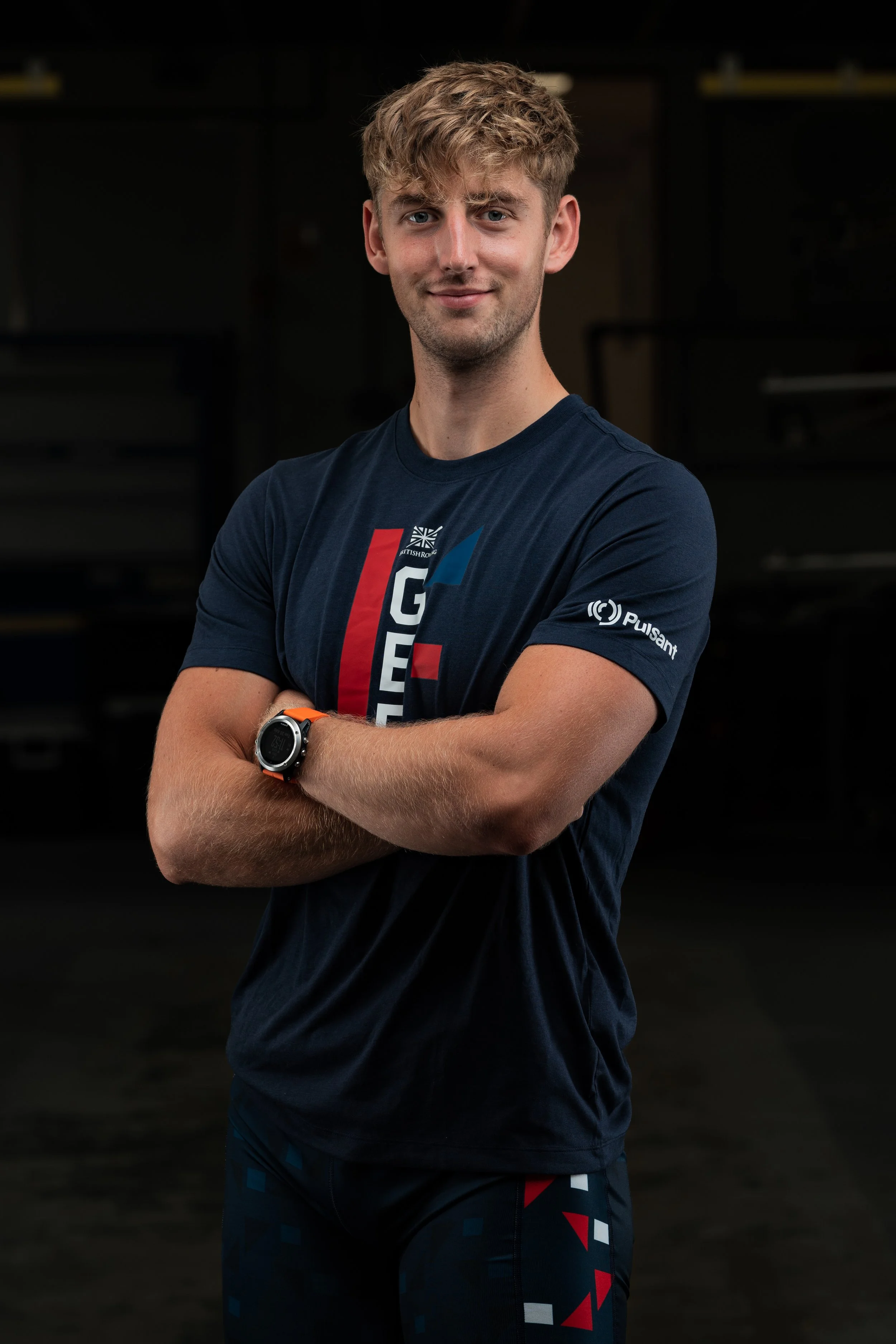 A young man with light brown curly hair and blue eyes standing with his arms crossed in a dark indoor gym or fitness center, wearing a navy blue athletic T-shirt and shorts, and a smartwatch on his left wrist.