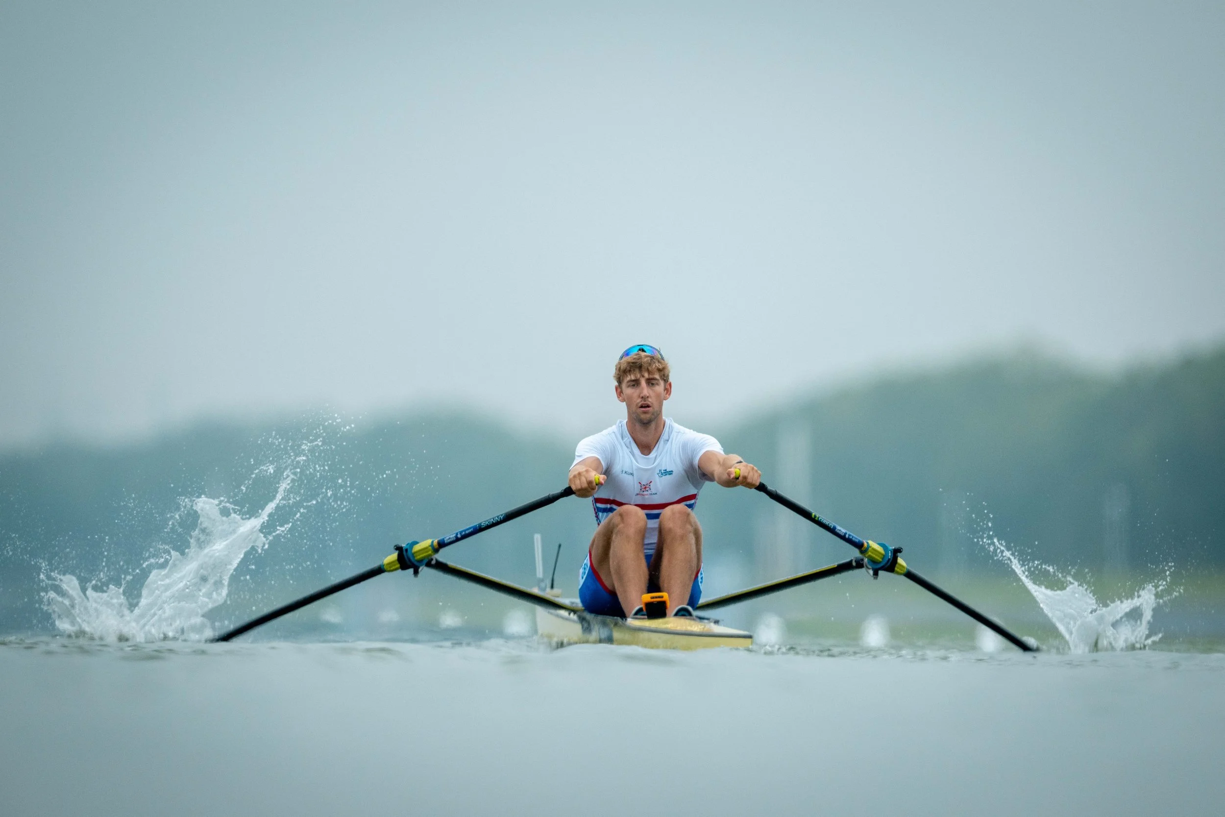 A male athlete is rowing a boat on a calm body of water, wearing a white athletic shirt with red and blue accents, a blue headband, and sunglasses. The sky in the background is overcast.