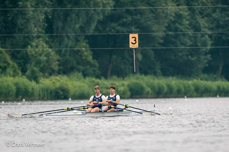 Two young men rowing a double scull boat on a lake with green trees in the background, a yellow sign with a black number 3 indicating water depth nearby.