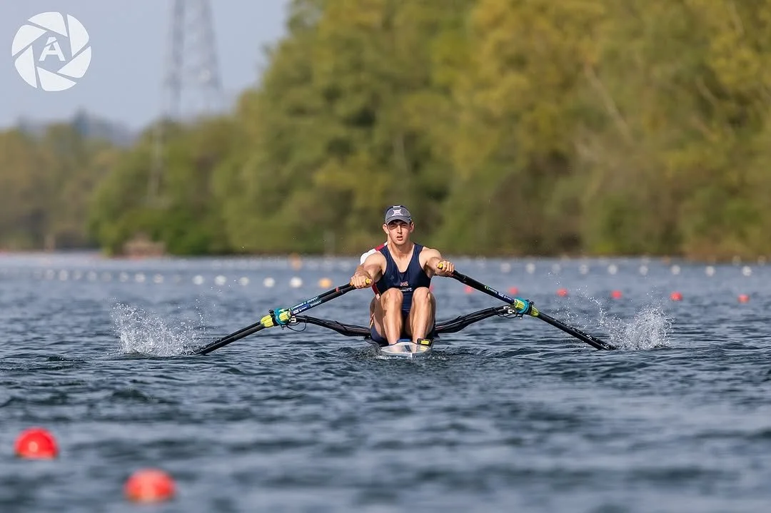 A male athlete rowing a single scull on a calm river with trees in the background during daytime.