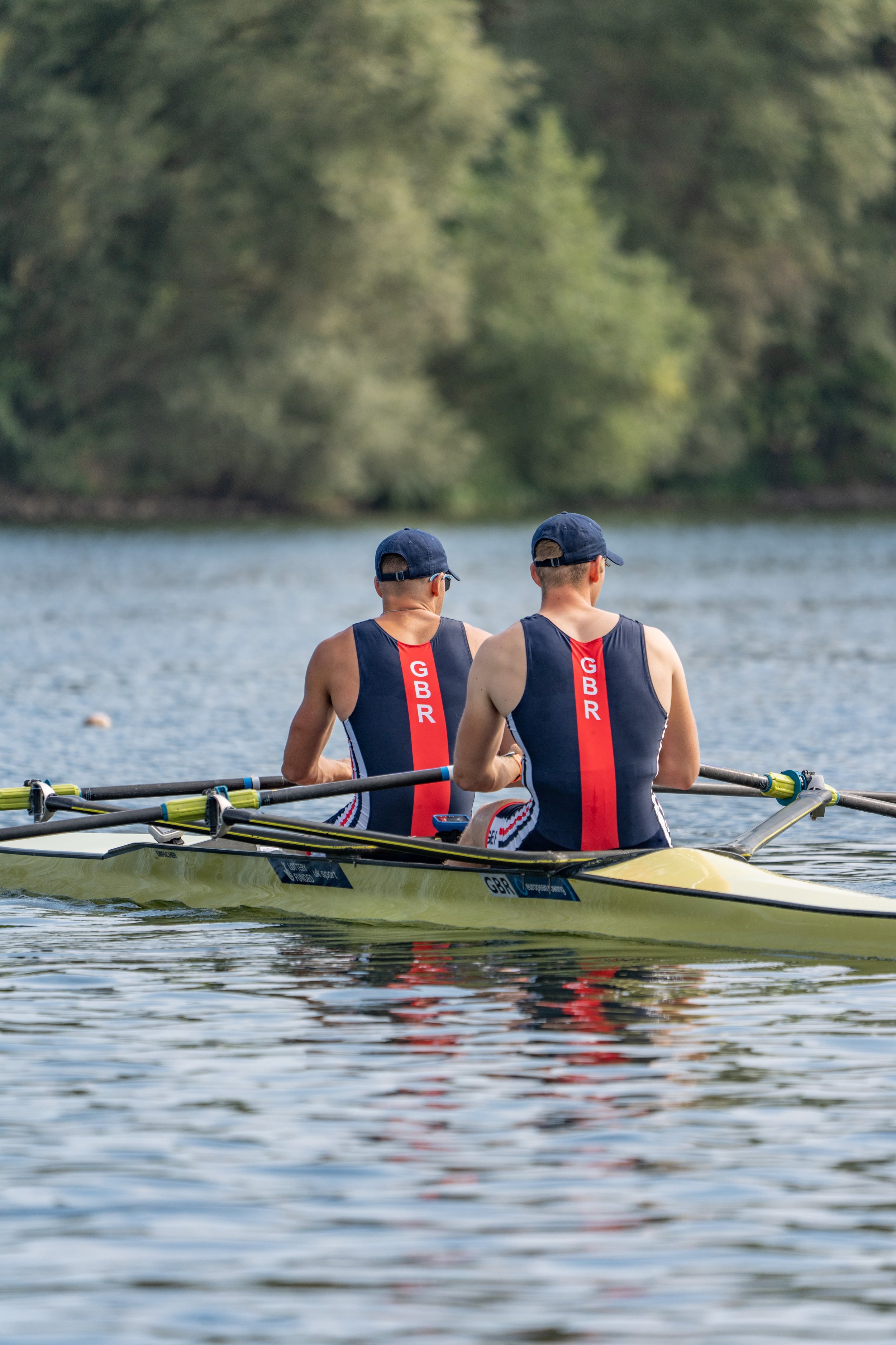Two rowers on a boat wearing matching navy blue and red uniforms with 'GBR' on the back, on a body of water with trees in the background.