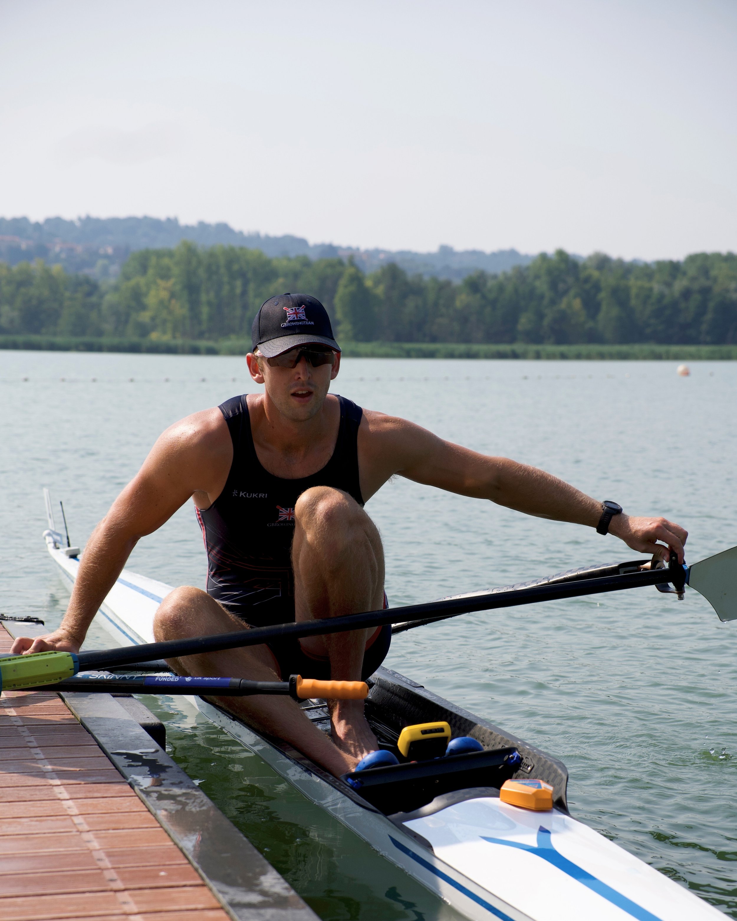 A male athlete dressed in a black athletic outfit and cap, sitting in a racing boat on a calm lake, with a green forested shoreline and hills in the background.