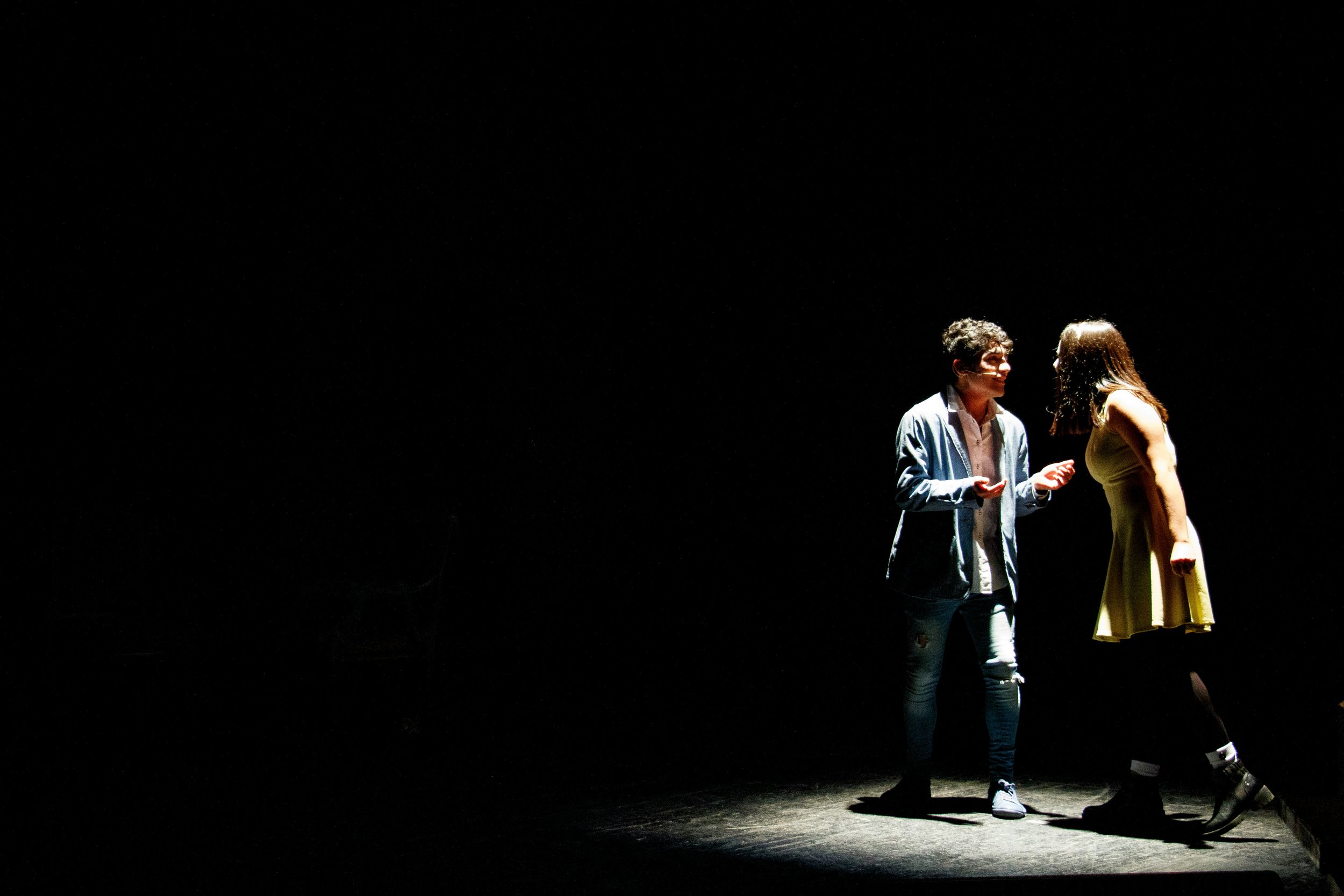 Two actors performing on a dark stage, illuminated from below. The male actor is wearing a light blue blazer and jeans, and the female actor is in a yellow dress and black boots. They are facing each other, engaging in dialogue.