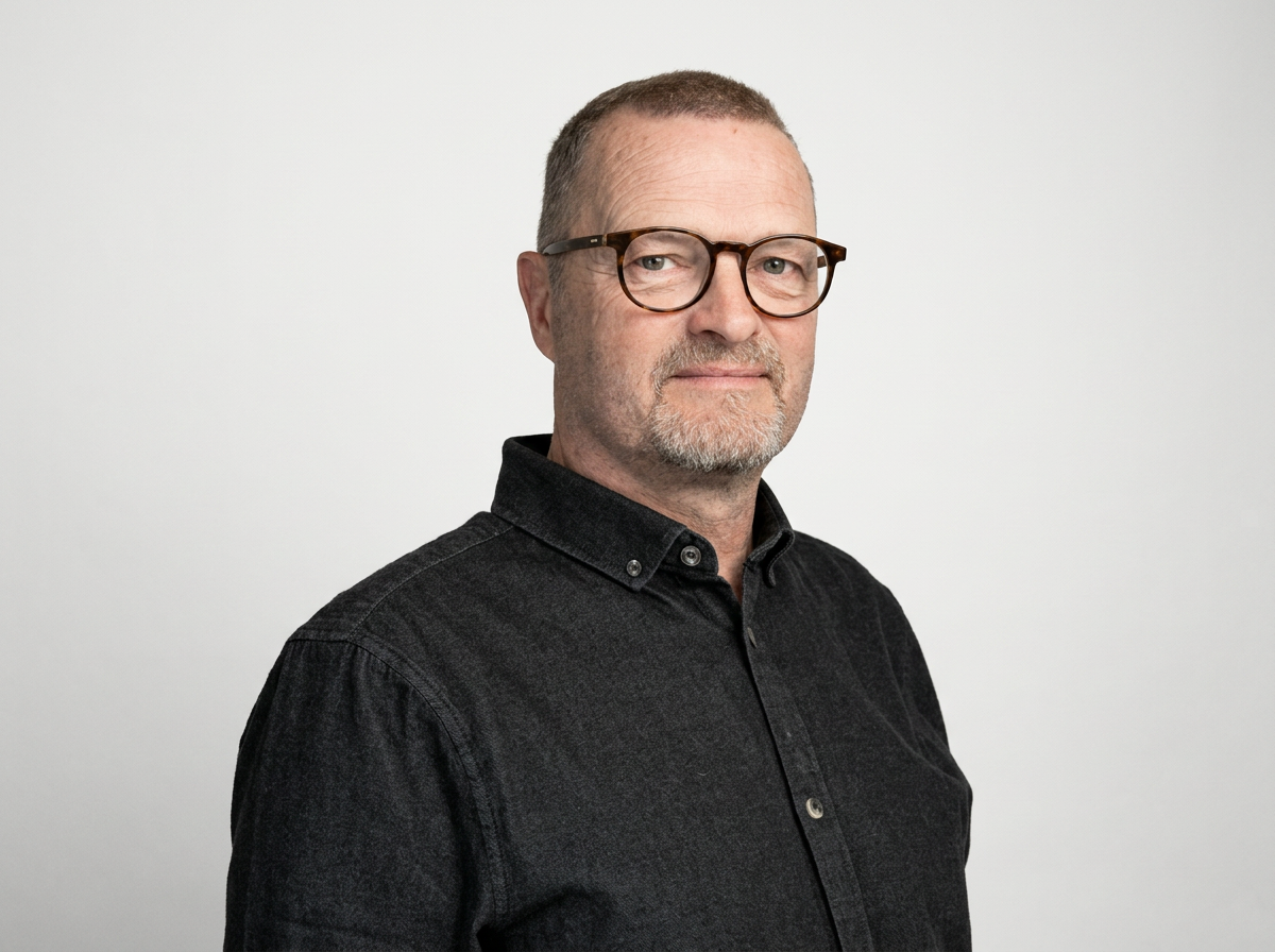 A middle-aged man with short hair and a goatee wearing glasses and a black button-up shirt, standing against a plain white background.