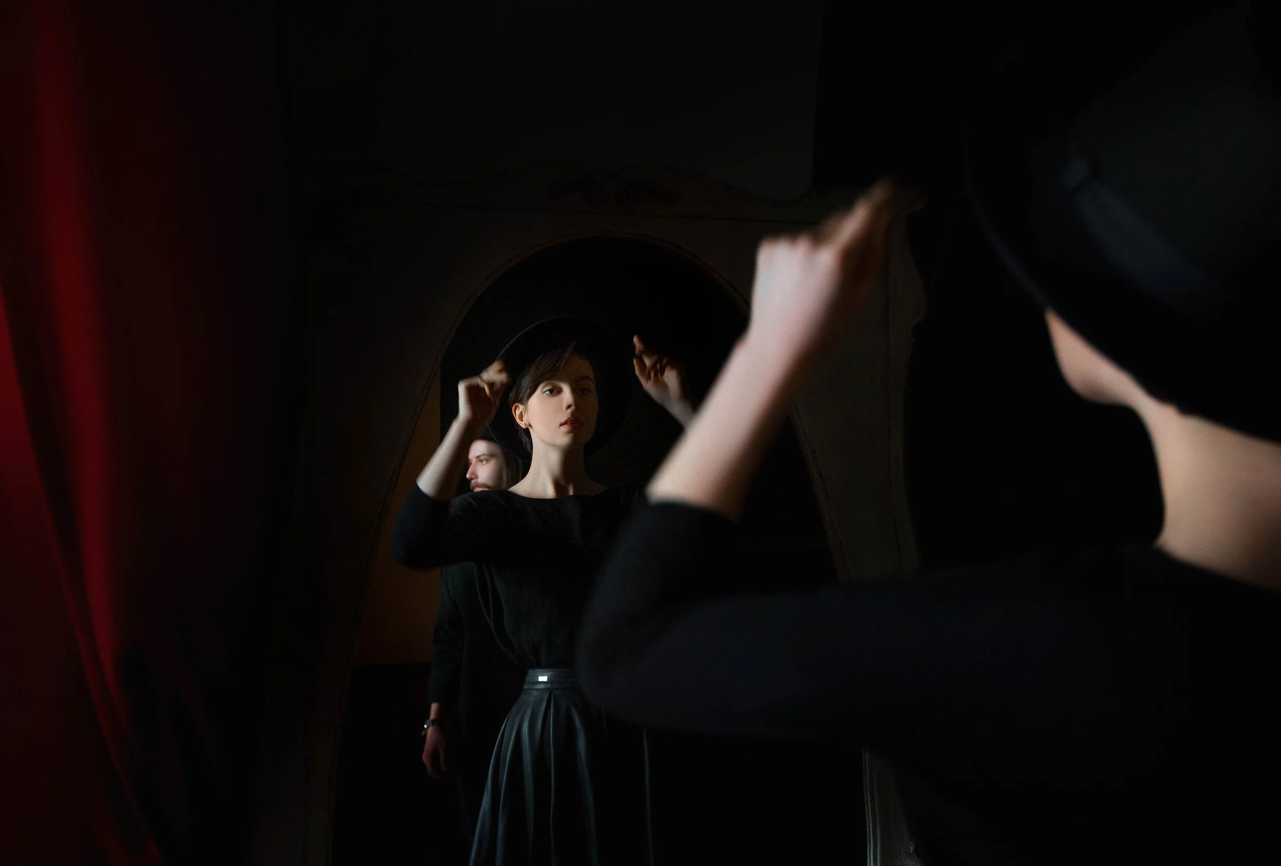 A woman standing in front of a mirror, adjusting her hat, in a dimly lit room.