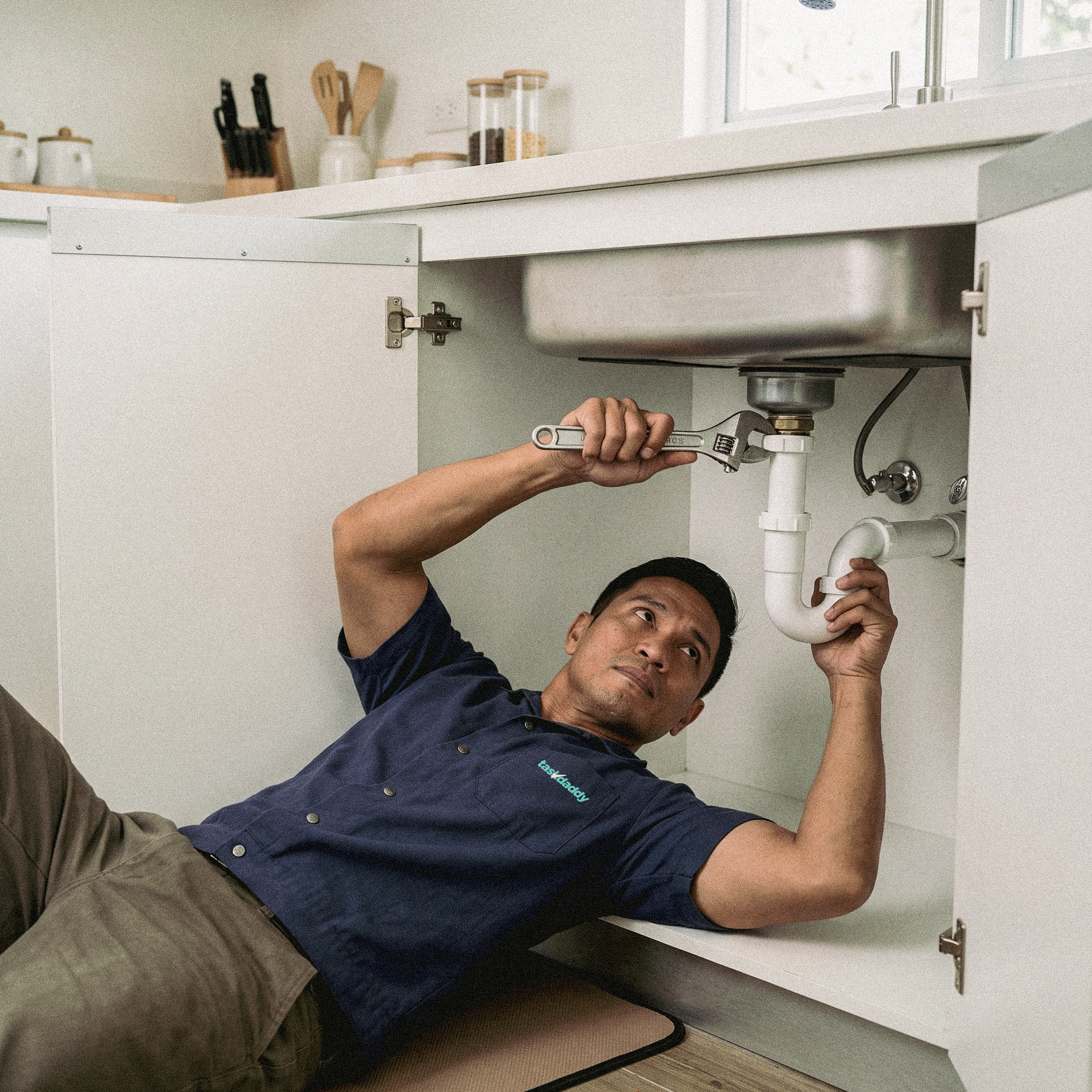 A technician working under a kitchen sink with a wrench, installing or repairing plumbing pipes, while lying on the floor.